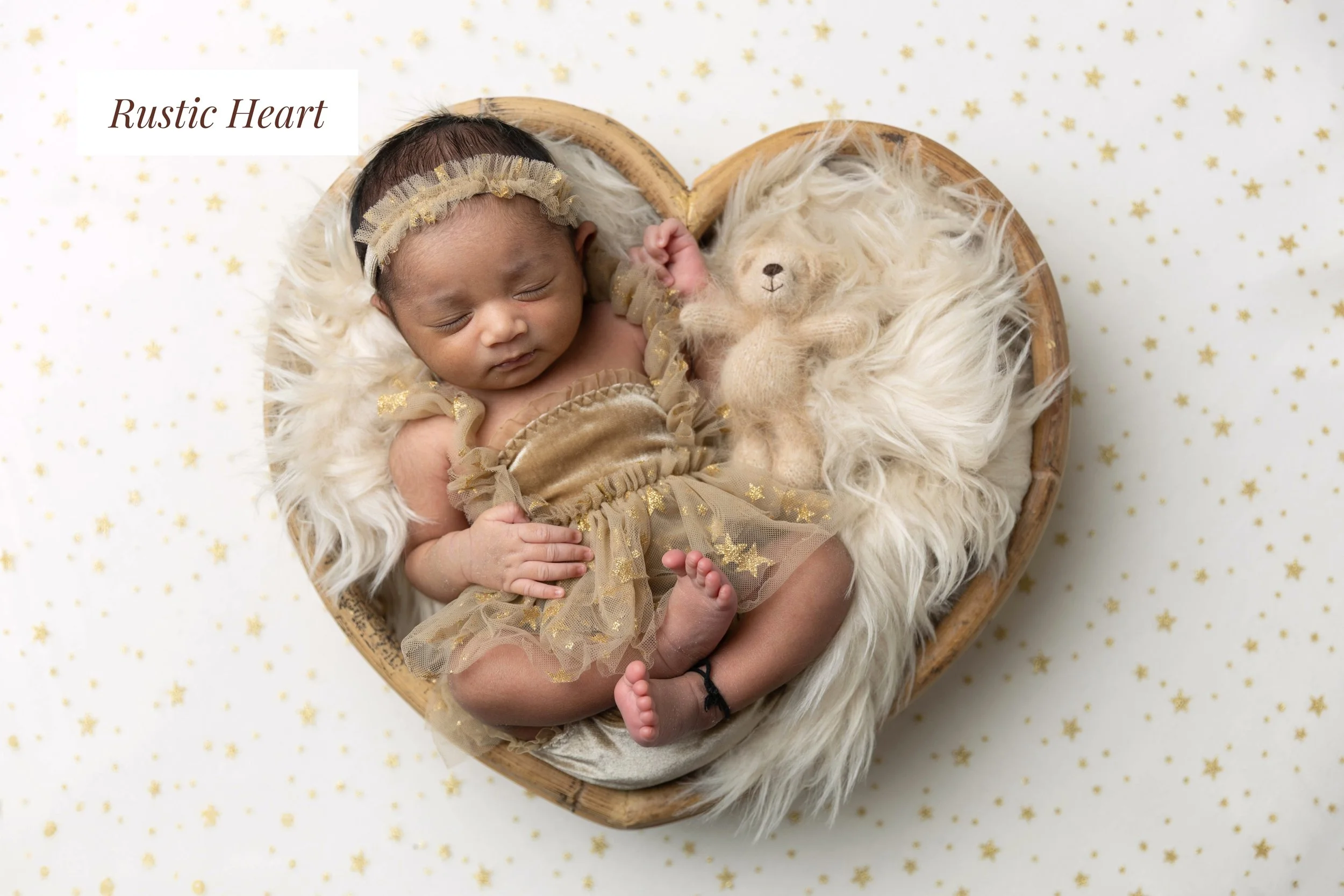 A newborn baby with a headband, sleeping on a fluffy white blanket in a heart-shaped wooden bed, next to a small teddy bear, with a white background decorated with small gold stars.