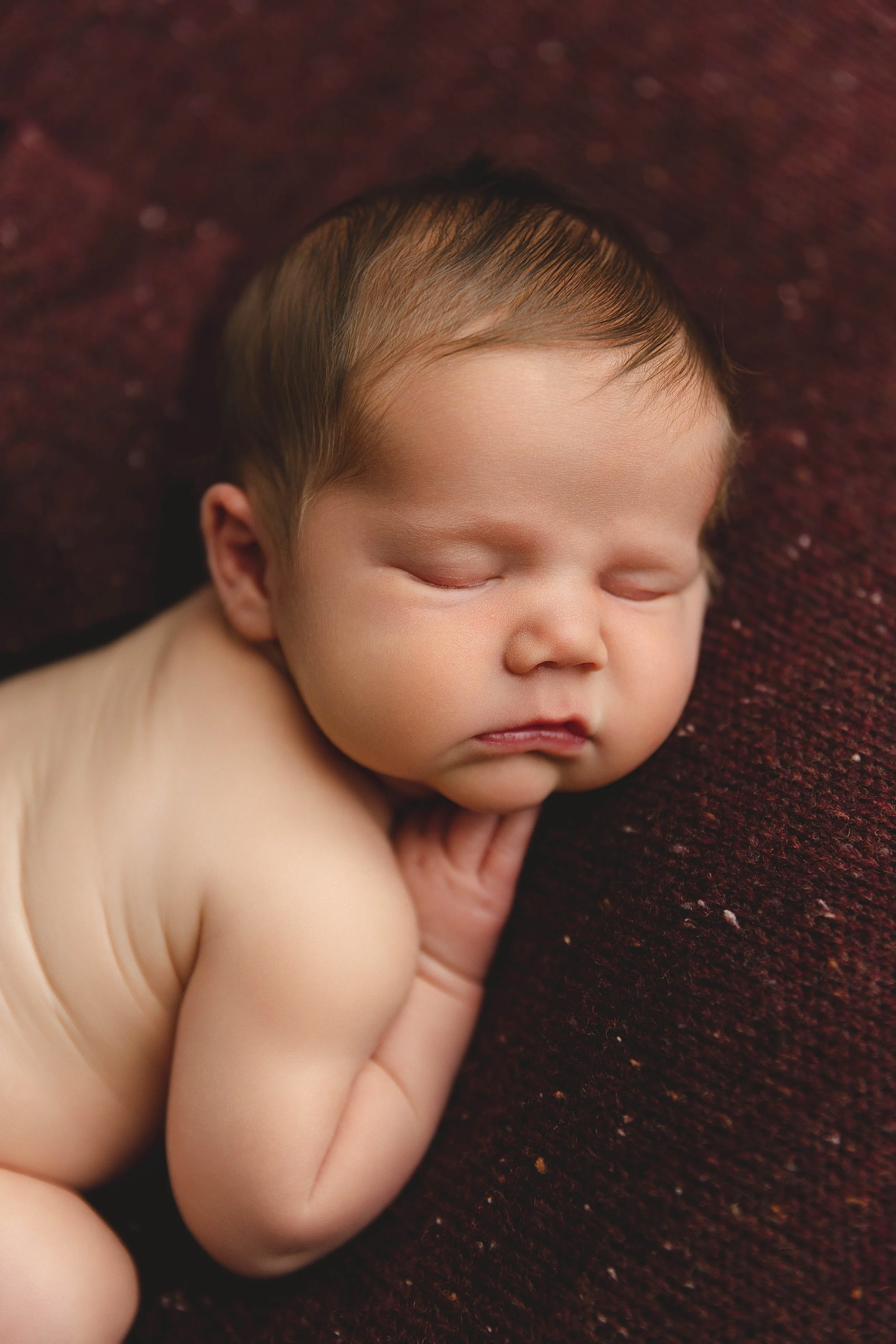 Close-up of a sleeping baby with fair skin and dark hair resting on a dark red textured surface.
