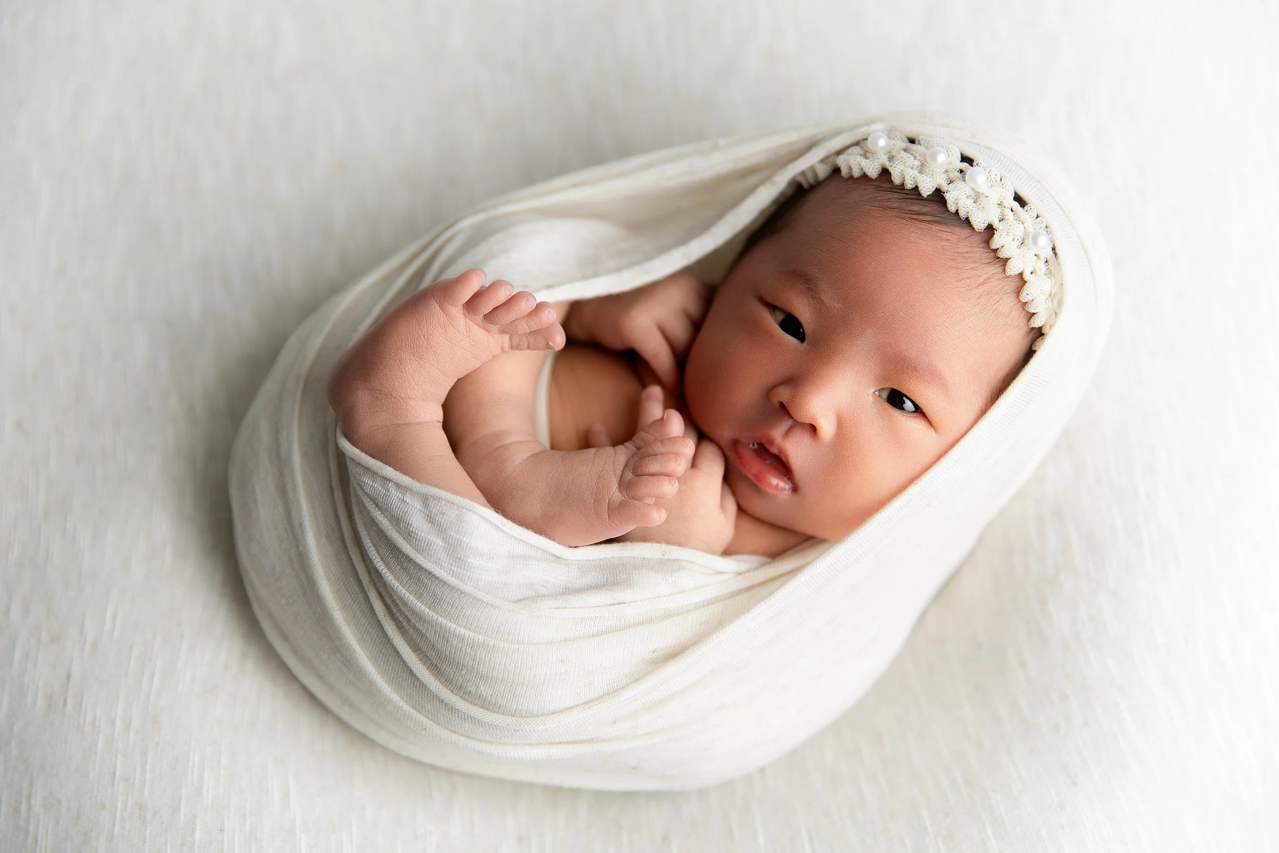 A newborn baby swaddled in a white blanket, wearing a headband with small flowers and pearls, lying on a white textured surface.