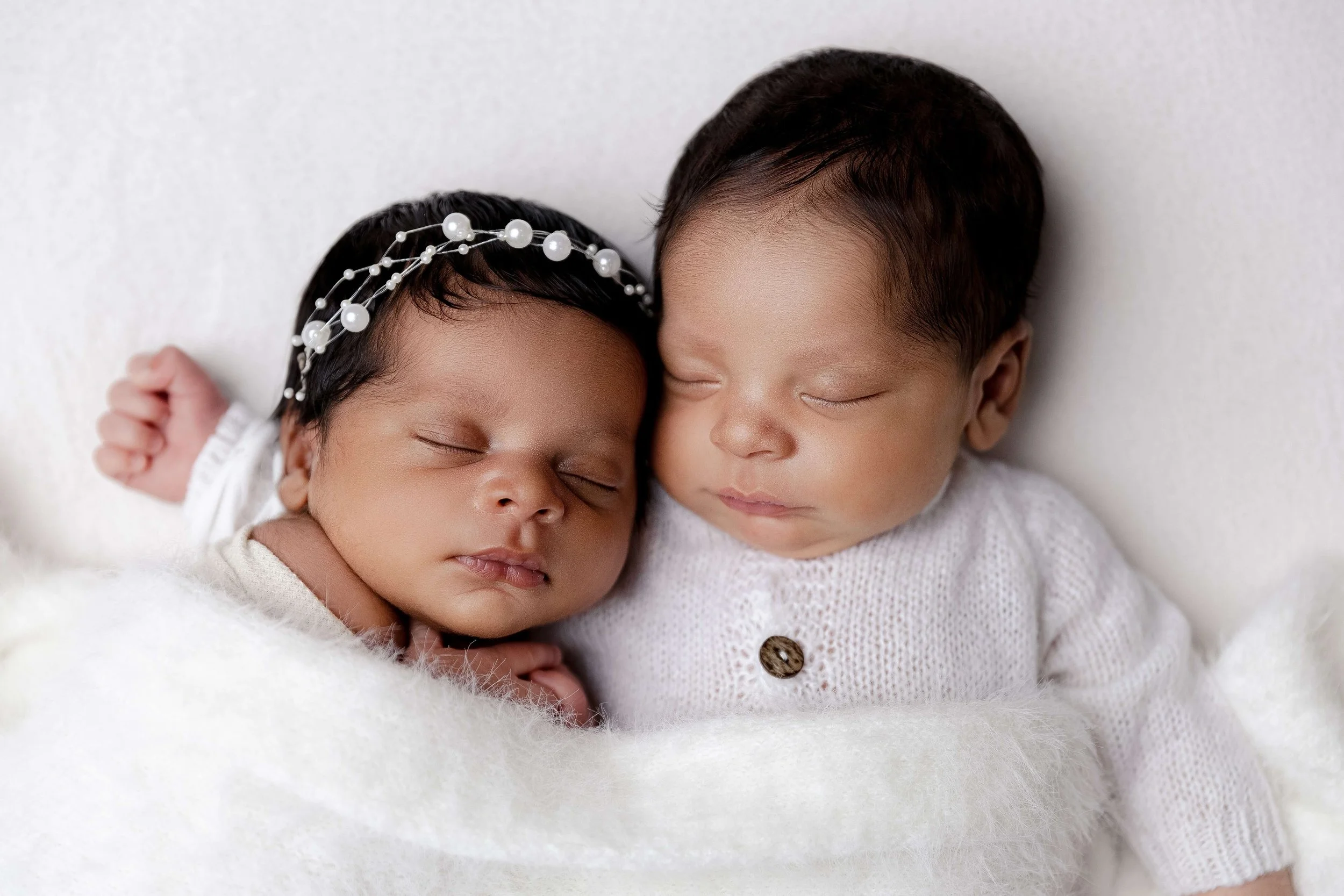 Two sleeping babies, one with a pearl headband, snuggling close on a white blanket.
