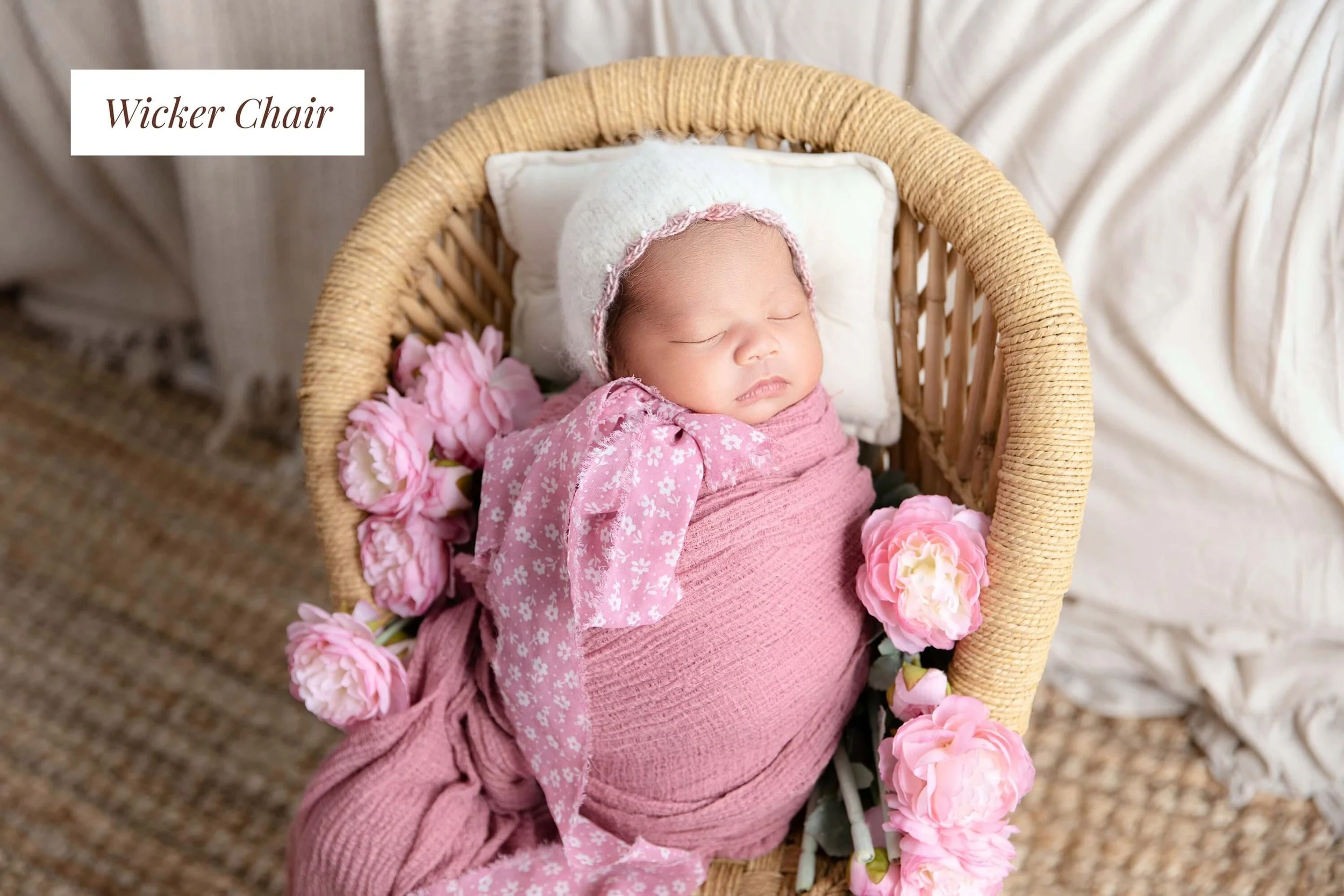A newborn baby girl sleeping in a wicker chair surrounded by pink flowers, wearing a pink floral outfit and a white knit hat.