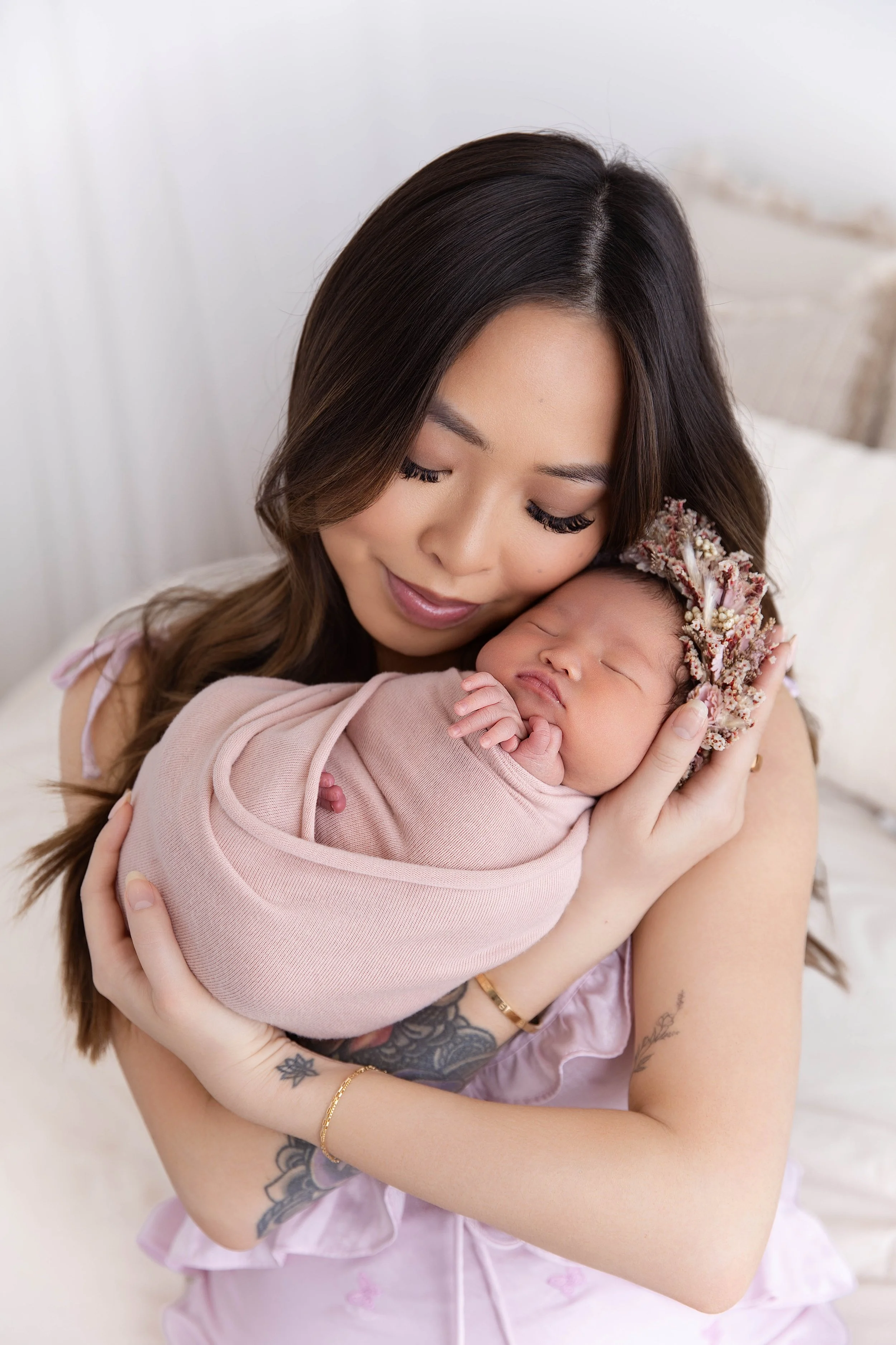 A woman holding a sleeping baby wrapped in pink clothing, wearing a floral headband, and sitting on a bed with light-colored bedding.