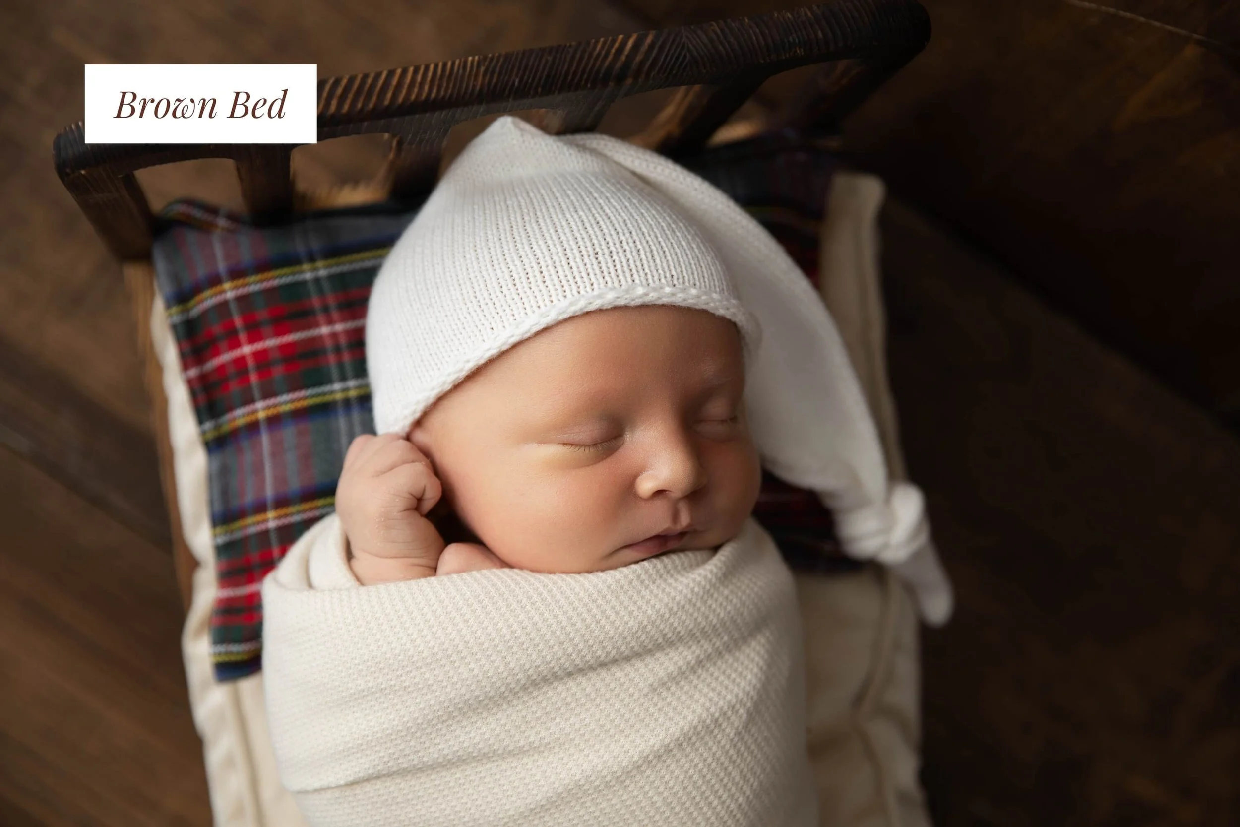 A sleeping baby wrapped in a cream blanket, wearing a white knit cap, resting in a brown wooden crib with a plaid pillow, on a wooden floor. The image has a label 'Brown Bed'.