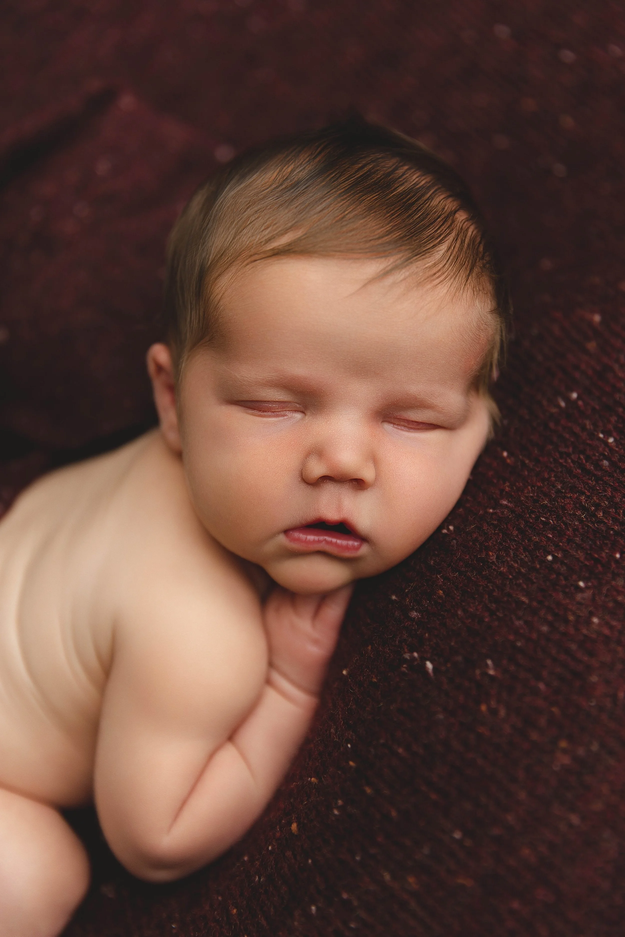 A sleeping baby with closed eyes and a slightly open mouth, lying on a textured dark red surface, supporting one cheek with a hand.