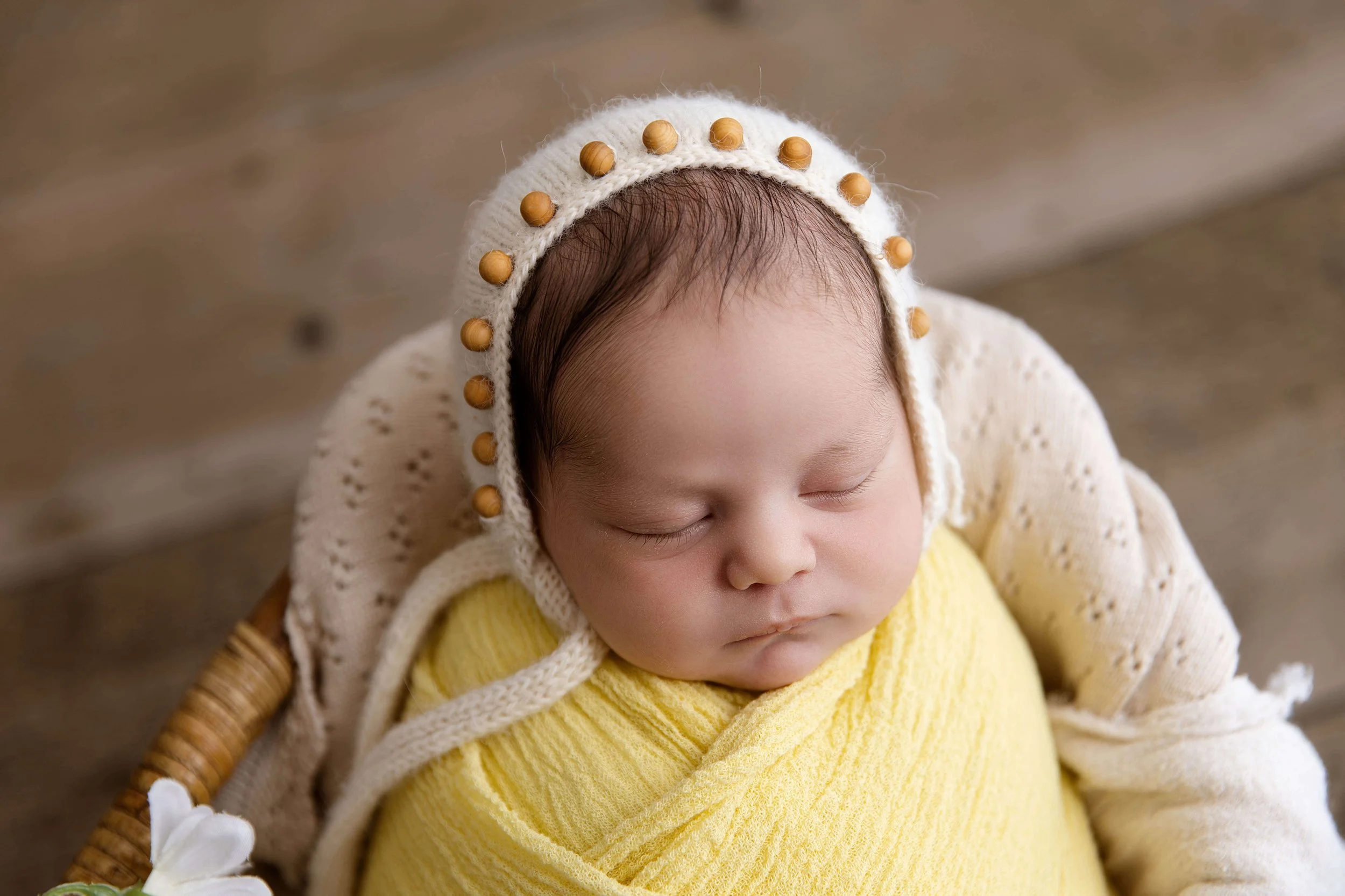 Close-up of a sleeping baby wearing a knitted cap with wooden beads and a yellow wrap, lying on a soft surface.