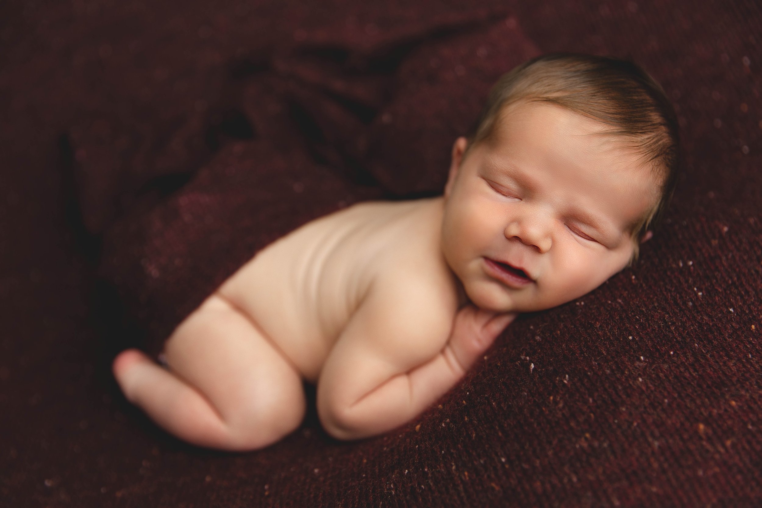 A sleeping newborn baby with dark hair, lying on a soft, dark maroon blanket, with a peaceful expression and arms tucked under their chin.