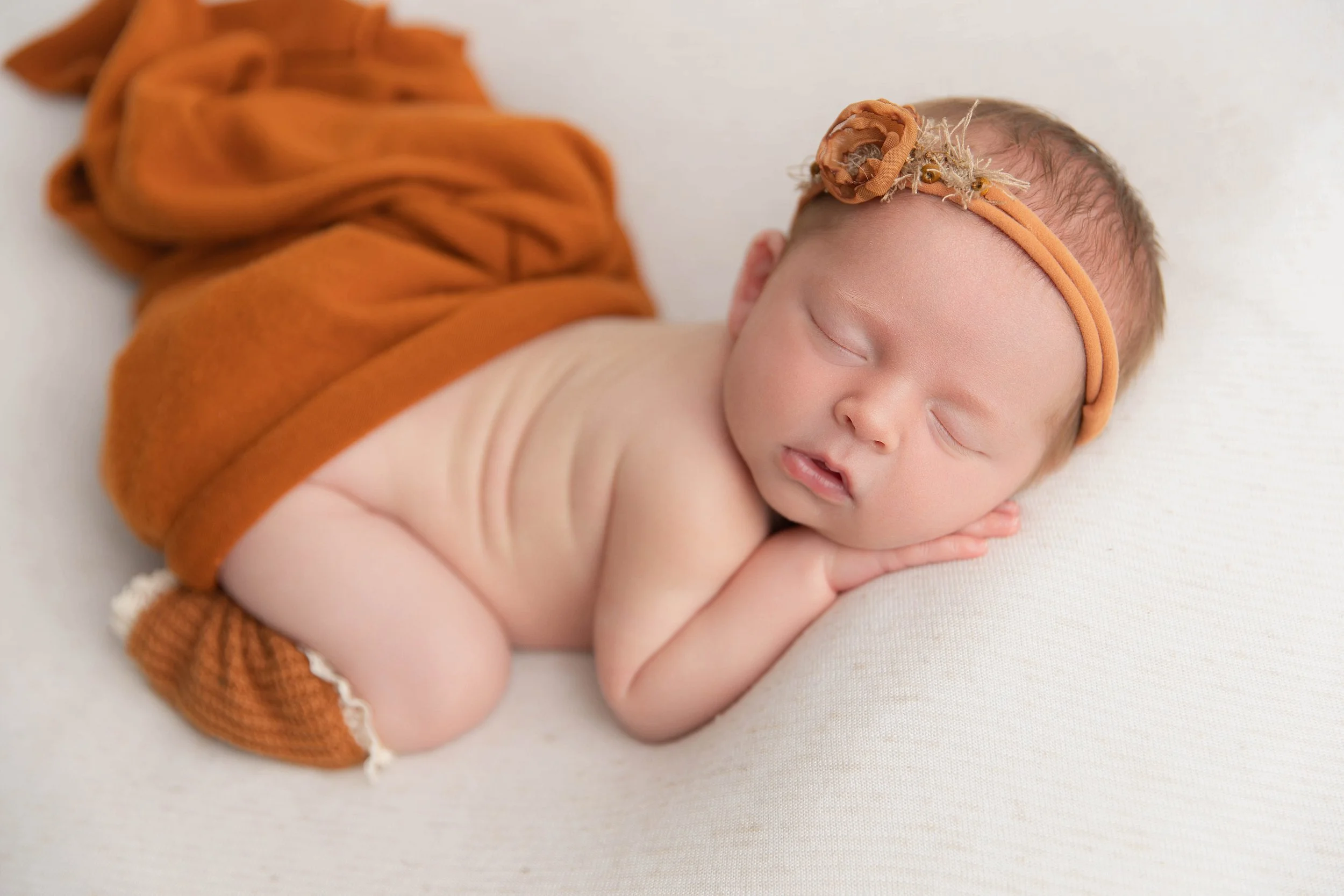 A sleeping newborn baby lying on a white surface, wearing a brown headband with a fabric flower, an orange wrap, and knitted socks.
