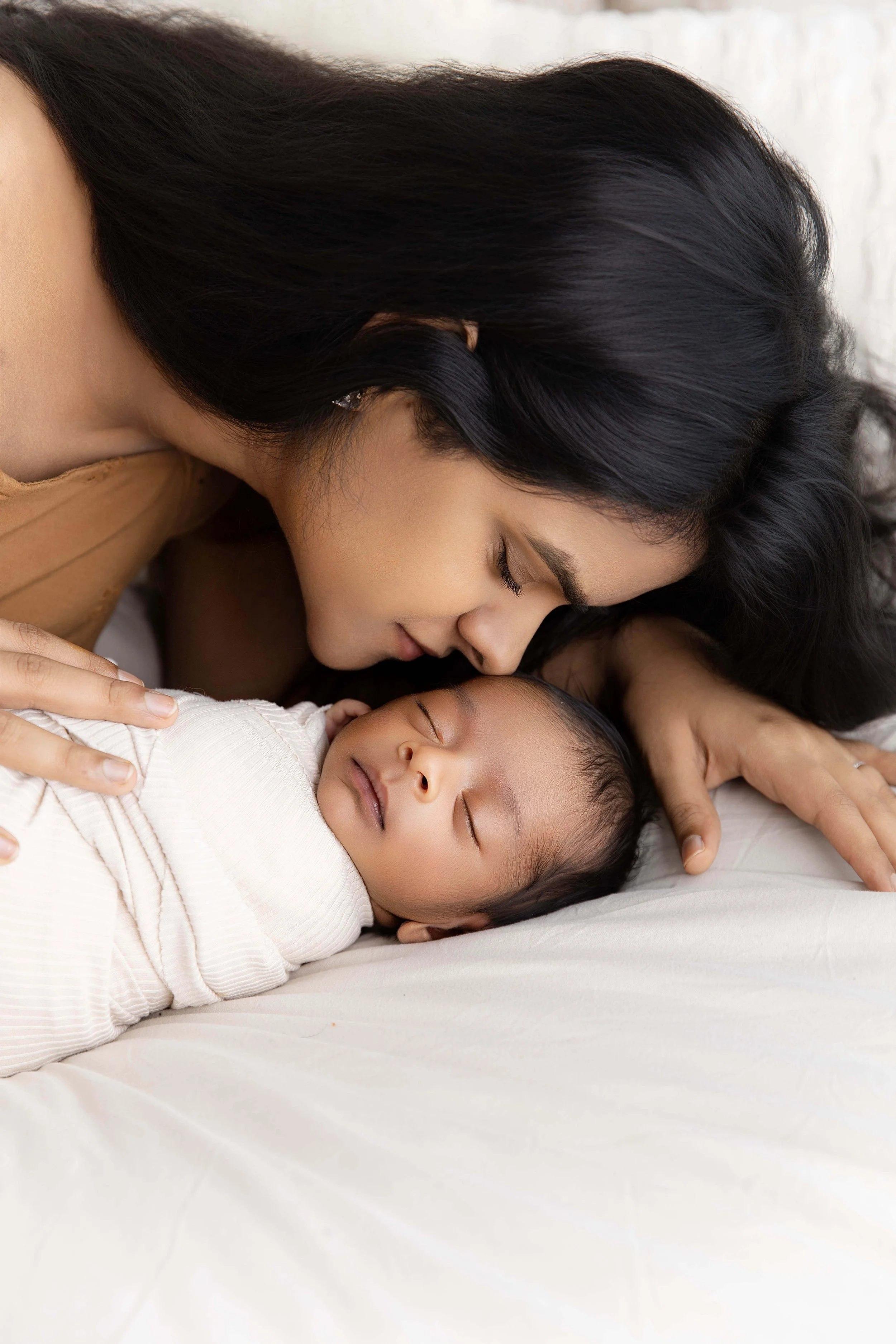 A woman and a baby lying on a bed, with the woman gently touching the baby and leaning in close with her eyes closed.