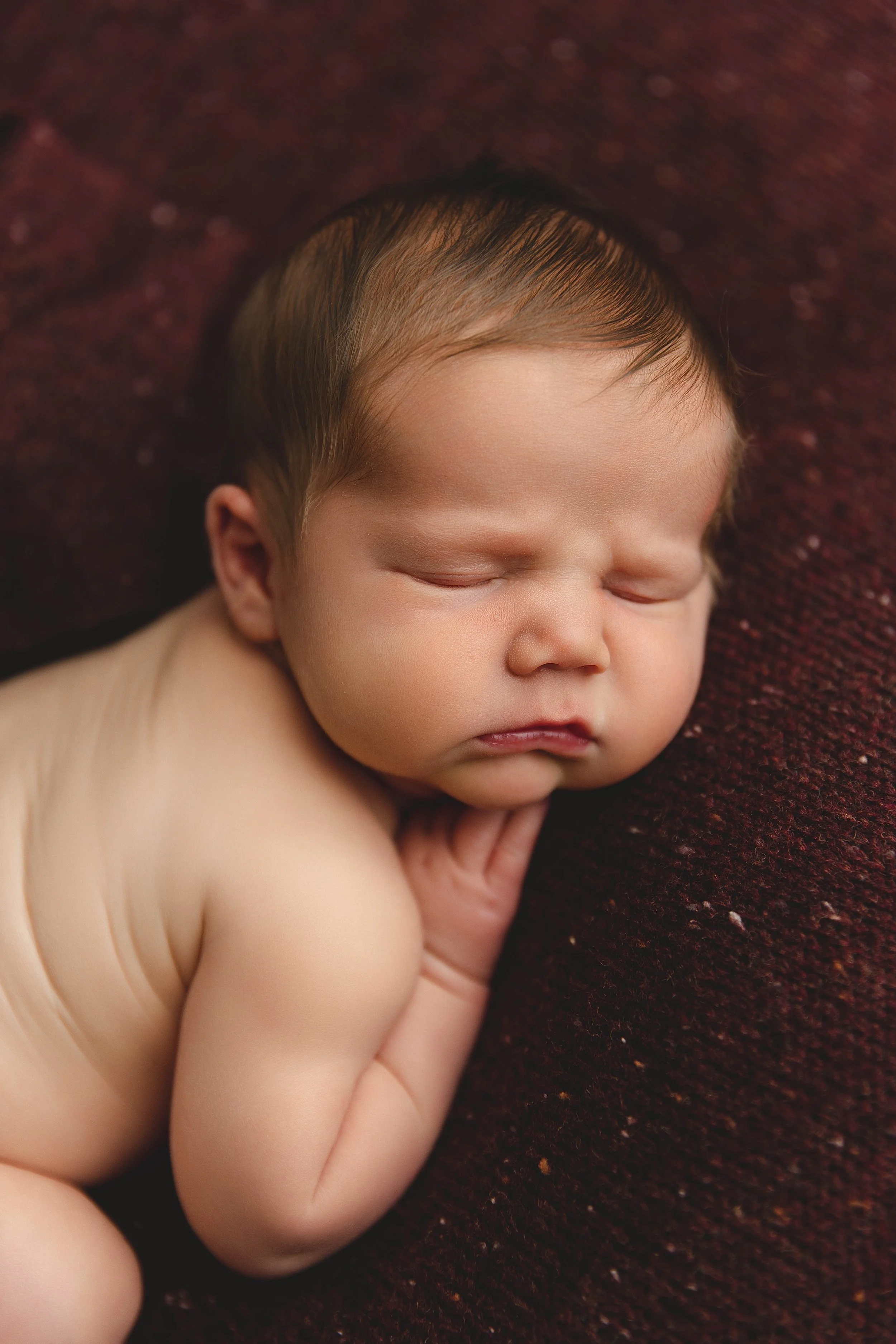 A sleeping newborn baby with fair skin and brown hair, lying on a dark red textured blanket.