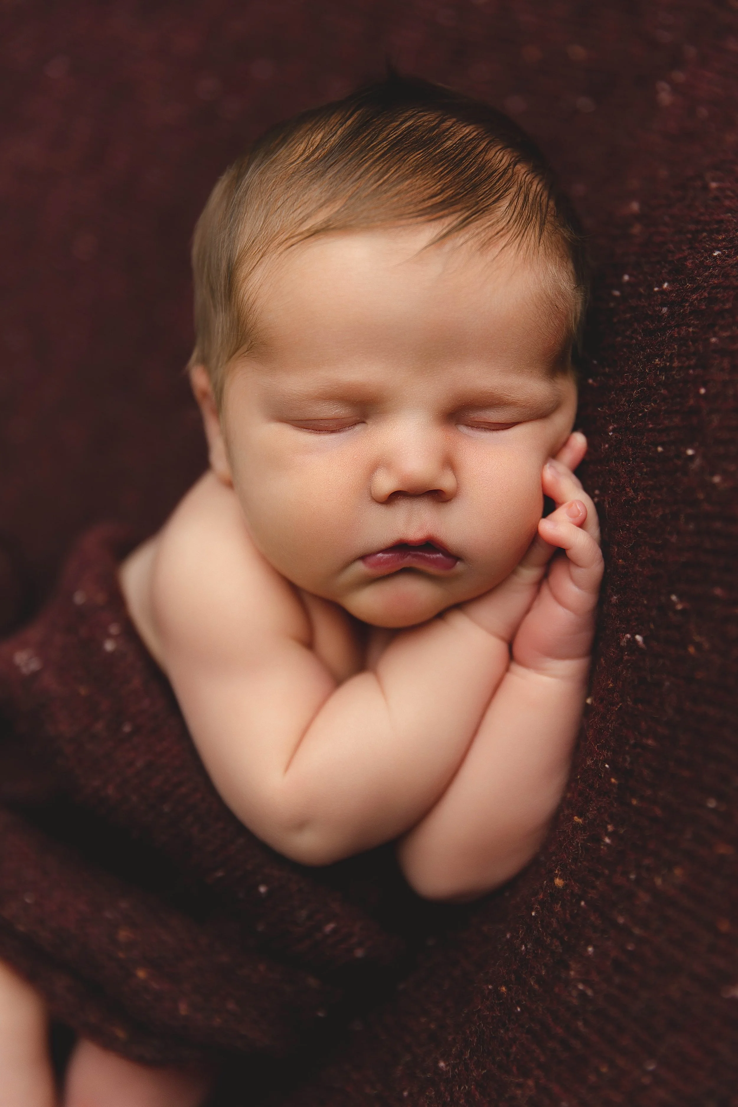 A sleeping newborn baby with closed eyes, resting its head on its hands, lying on a dark brown textured blanket.