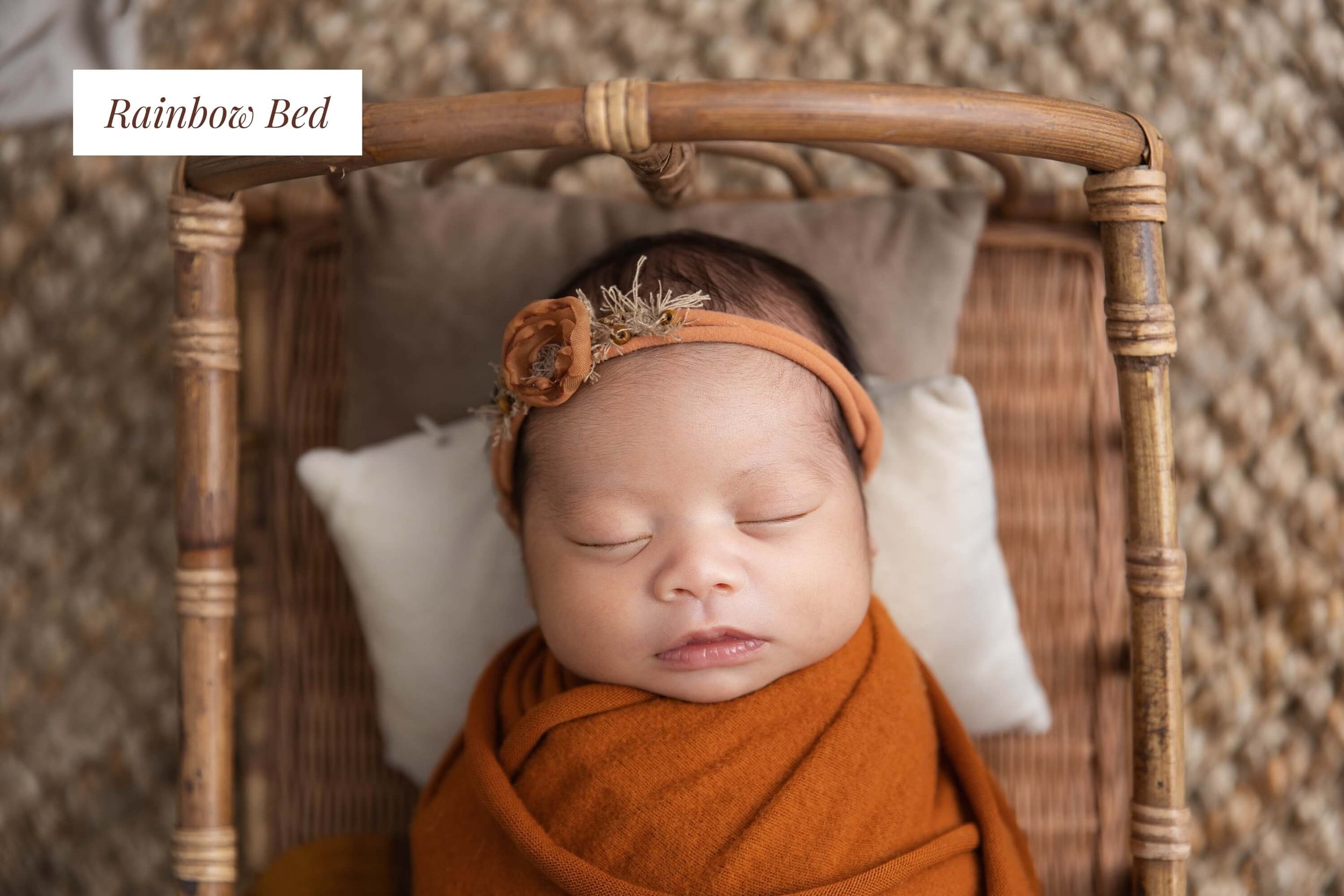 A sleeping baby lying on a small wicker bed with a pillow, dressed in a rust-colored outfit and a matching headband with a flower, on a textured surface.