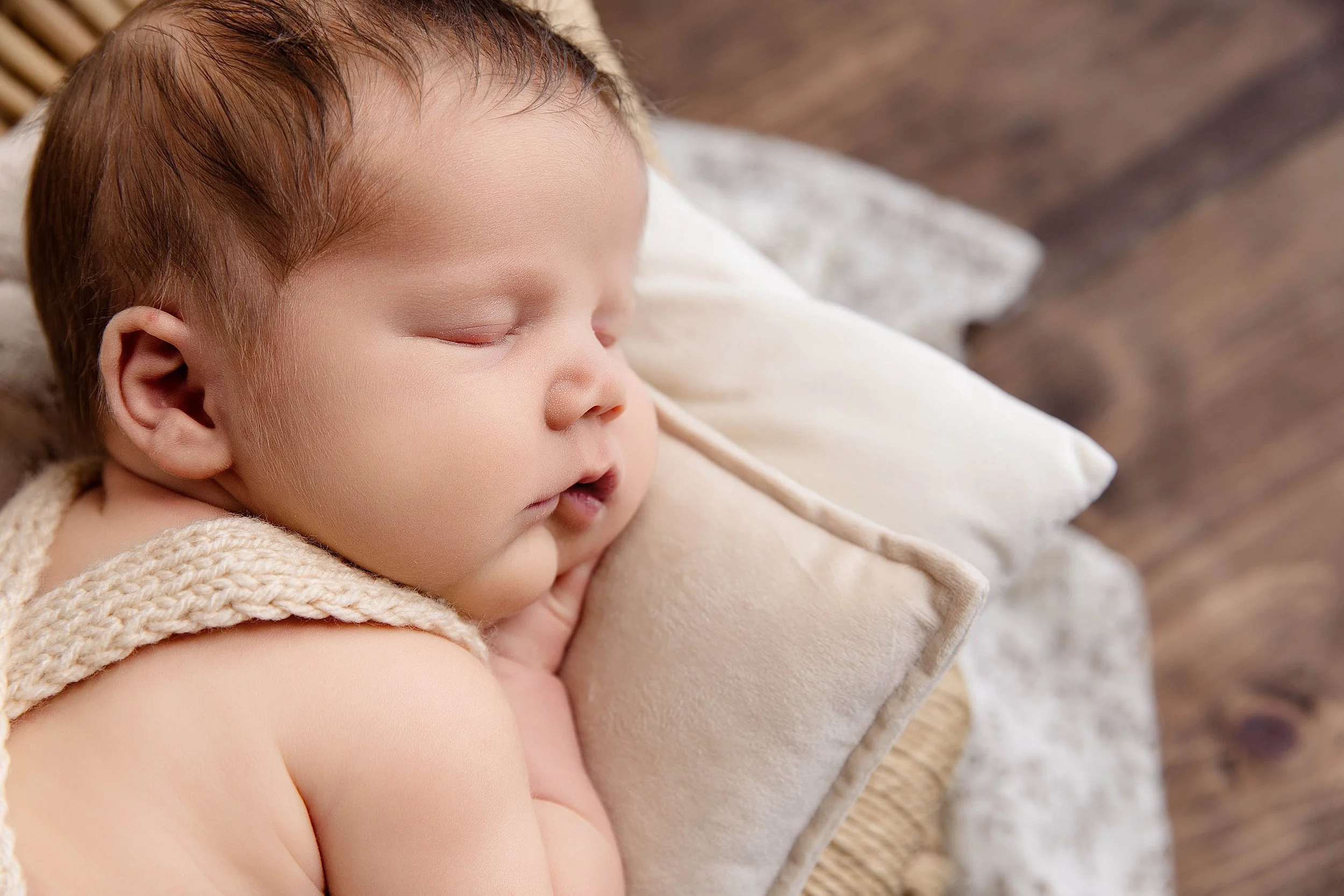 A close-up of a sleeping baby with light skin, brown hair, and closed eyes, resting on a beige pillow and covered with a knitted blanket.
