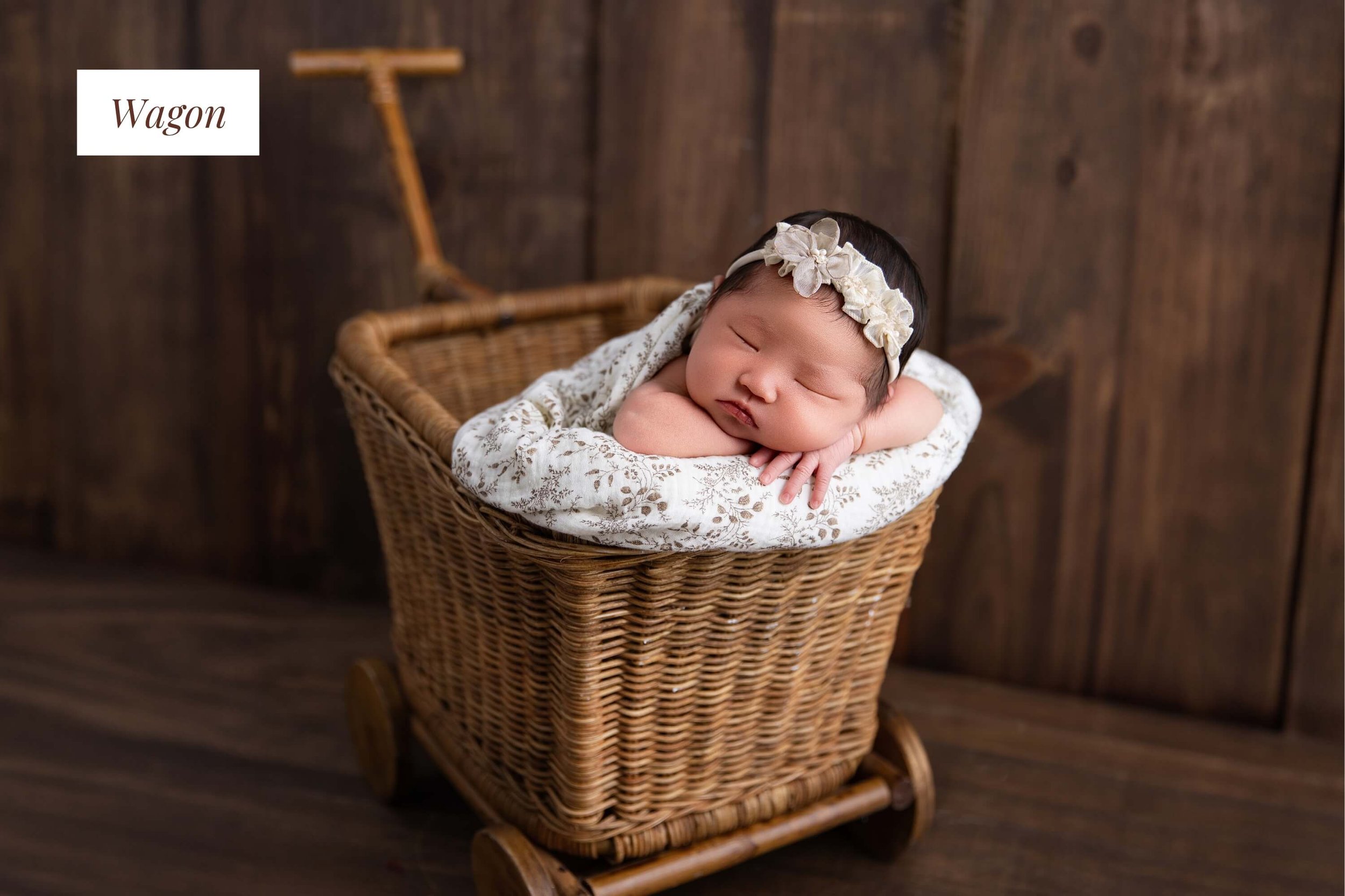 A sleeping baby girl with a floral headband, nestled in a wicker wagon with a patterned blanket, against a rustic wooden backdrop.