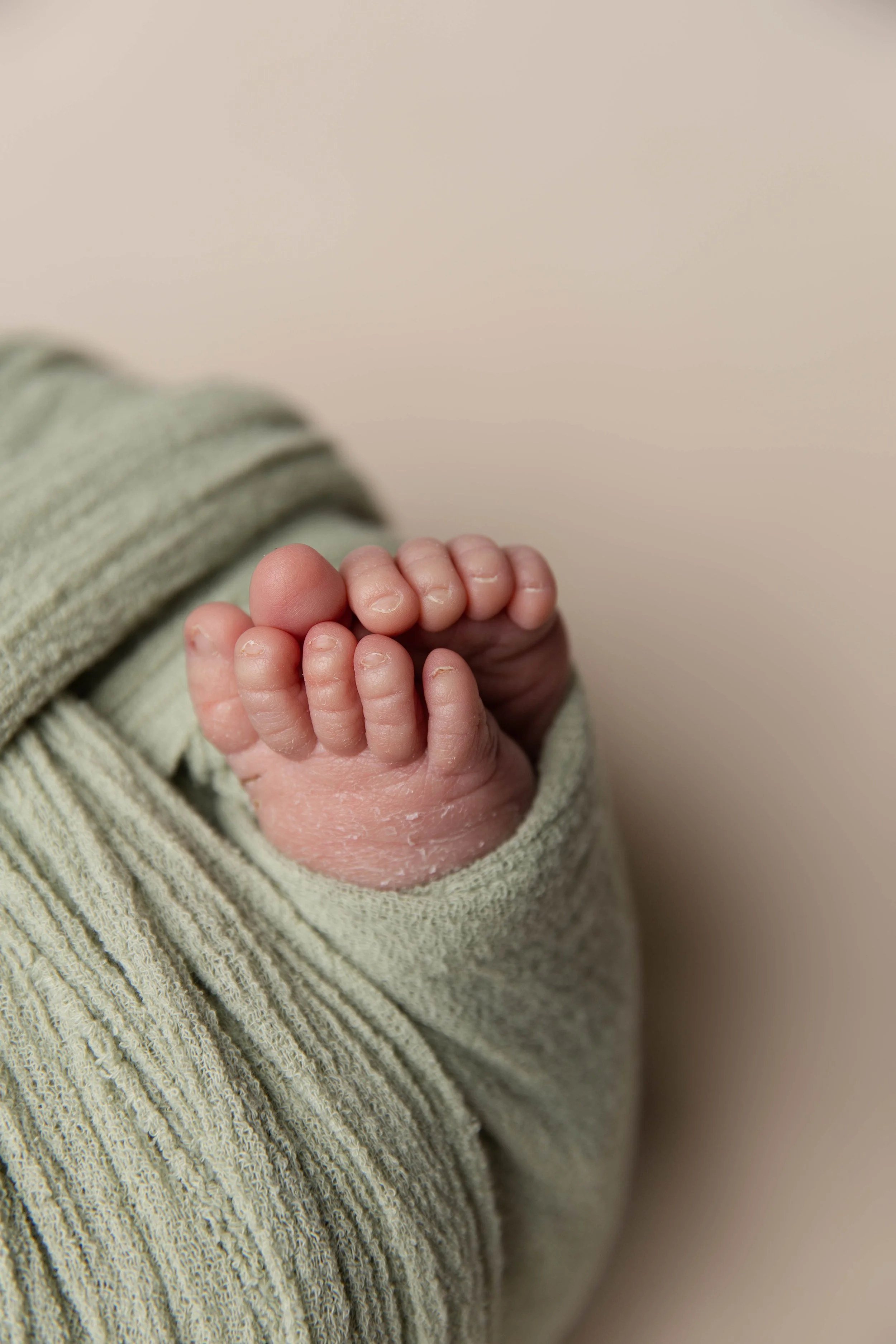 Close-up of a newborn baby's tiny hand curled into a fist, wrapped in a soft, green knitted blanket, against a neutral background.