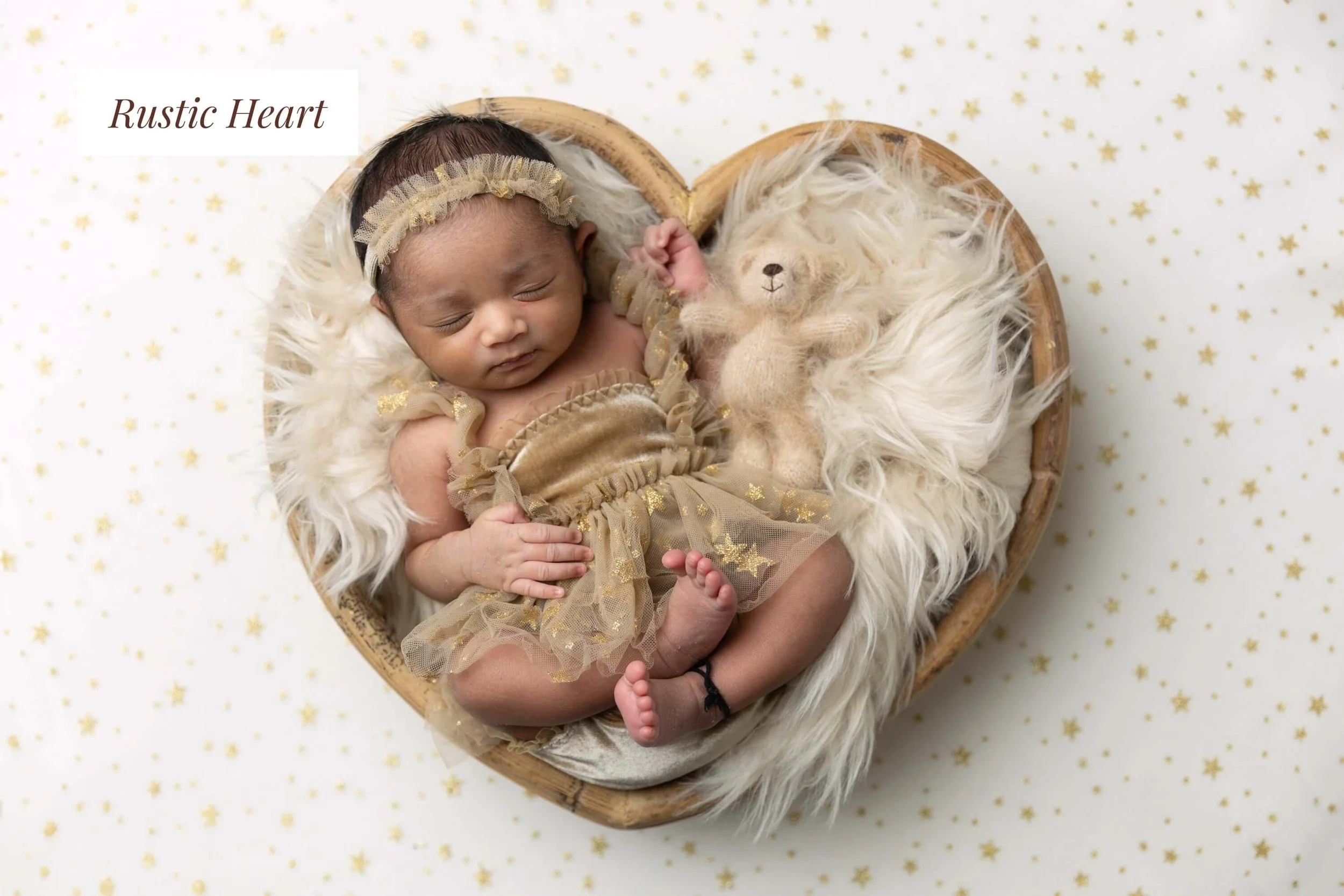 A sleeping baby in a gold outfit with a matching headband, lying in a heart-shaped wicker basket with a faux fur lining and a small plush bear, surrounded by a white background with gold star patterns.