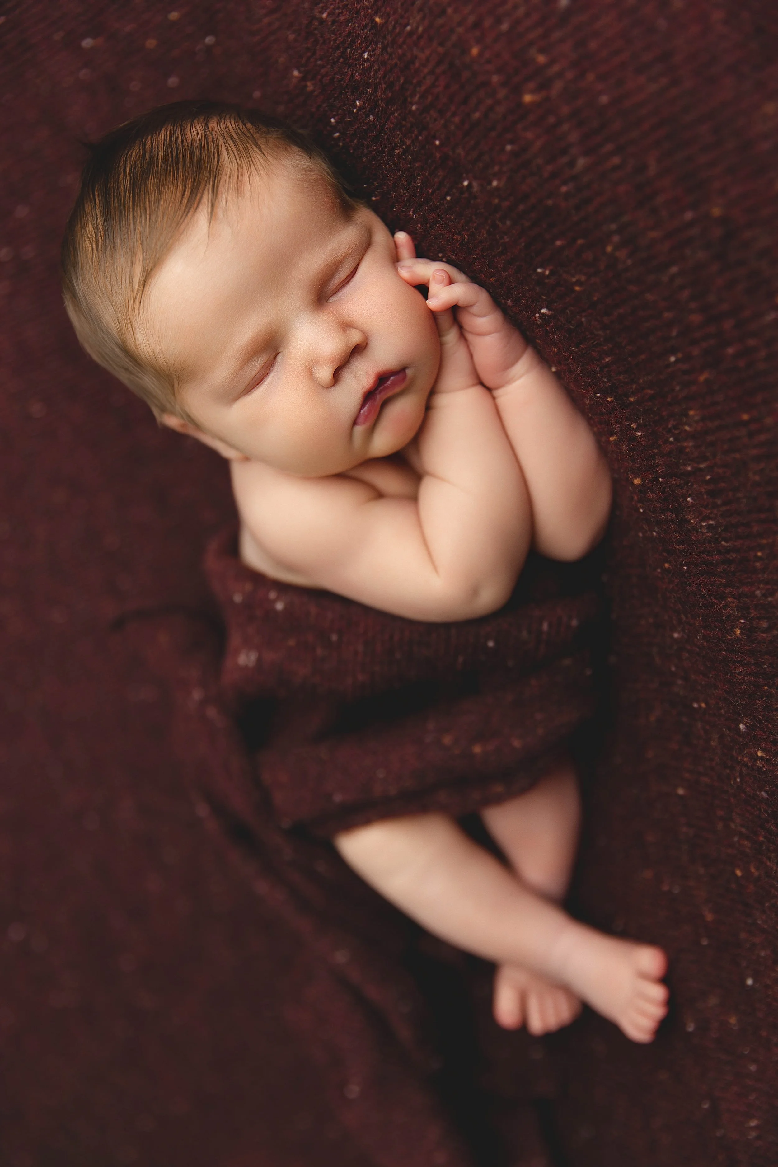 A sleeping baby with light skin, light brown hair, lying on a burgundy blanket, with eyes closed and hands tucked under the cheek.