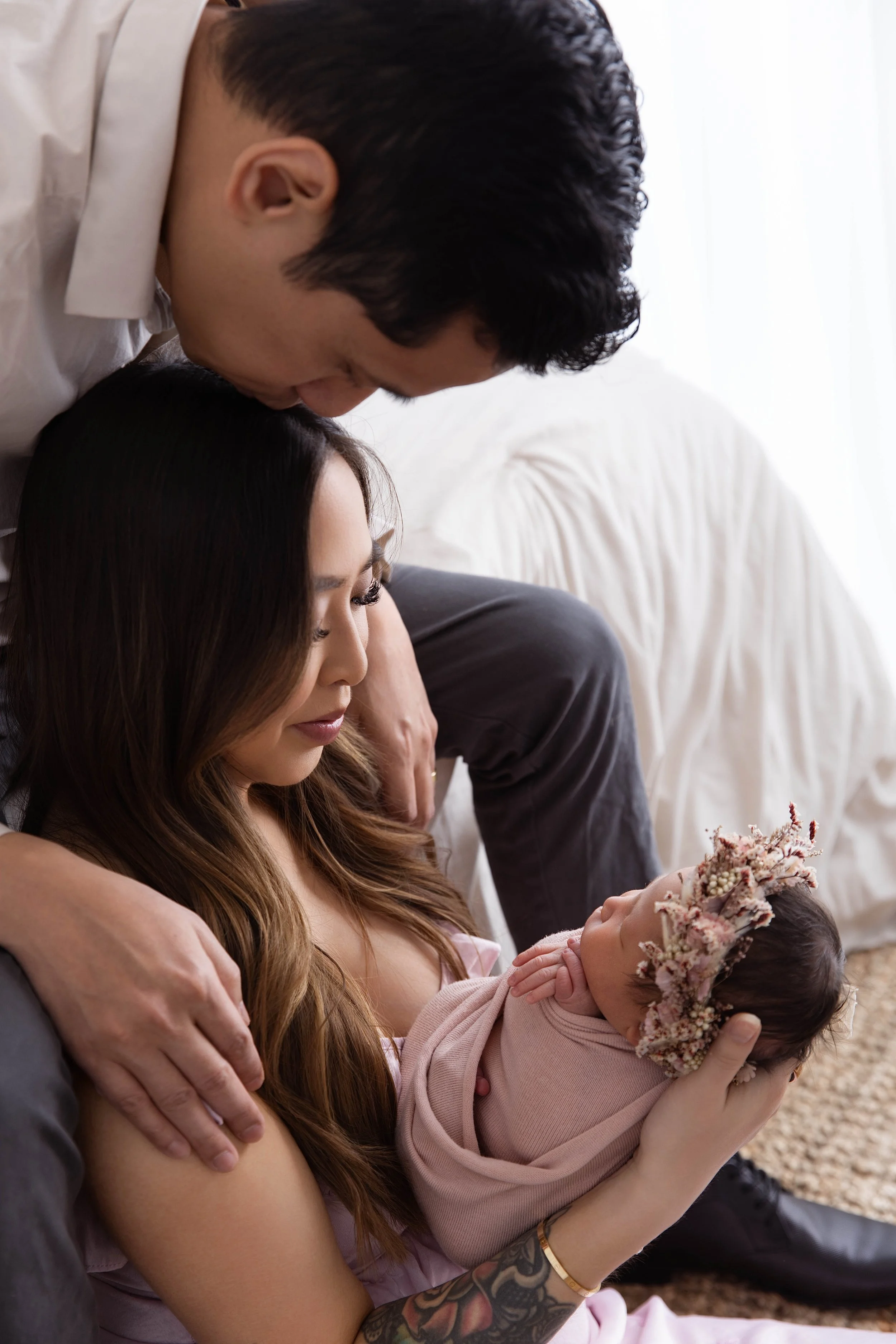 A woman with long brown hair holding a newborn baby wrapped in pink with a floral crown, while a man leans over and looks at the baby tenderly.