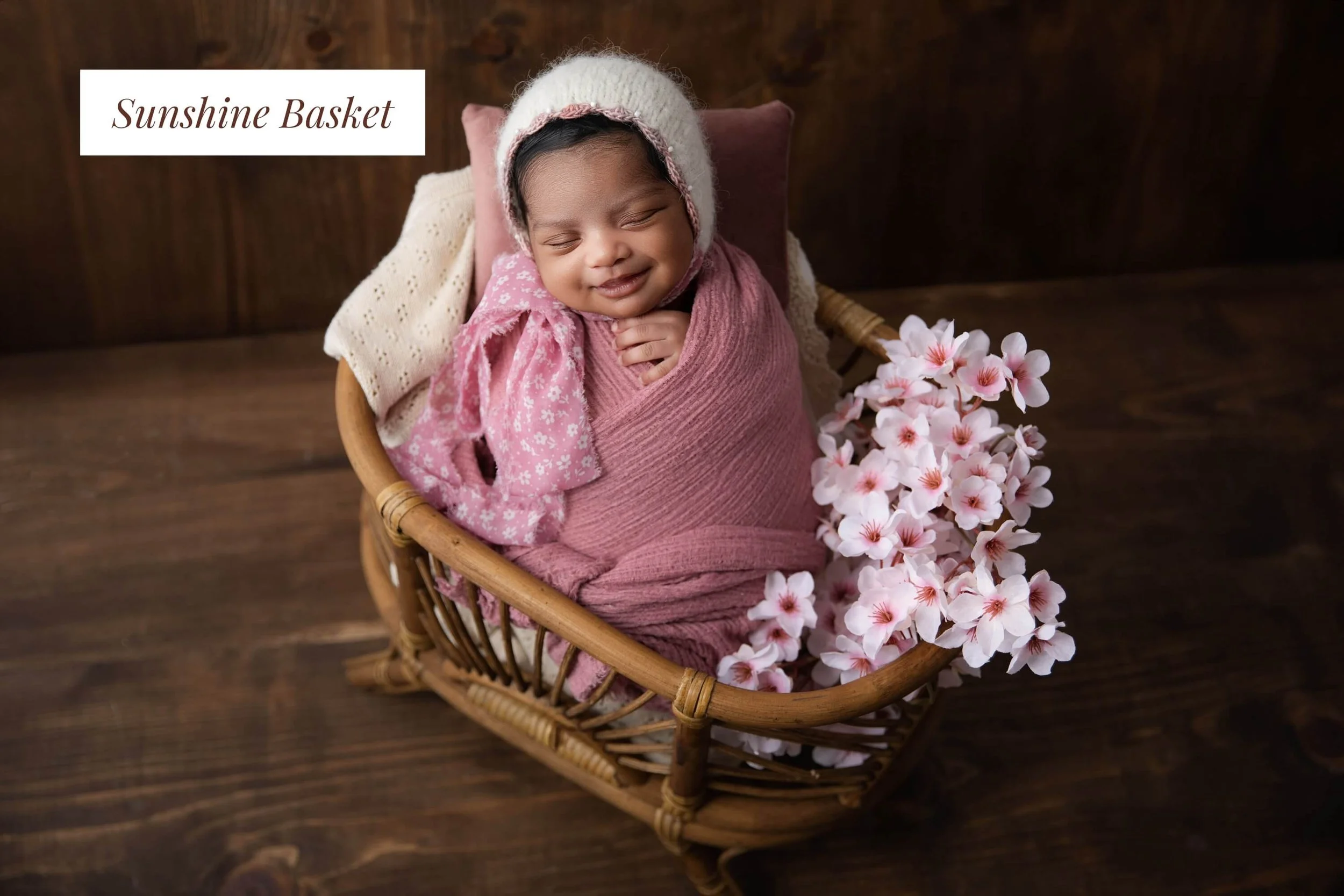 A smiling baby wrapped in pink fabric, wearing a white knit hat with pink trim, lying in a wicker basket filled with pink and white flowers on a dark wooden surface, with a white label that says "Sunshine Basket"