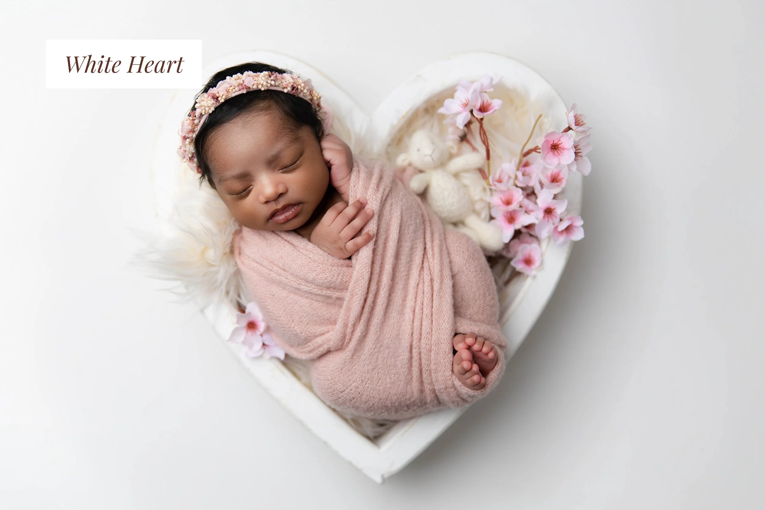 A sleeping newborn baby girl wrapped in pink cloth, lying in a white heart-shaped basket decorated with pink flowers and stuffed animals.