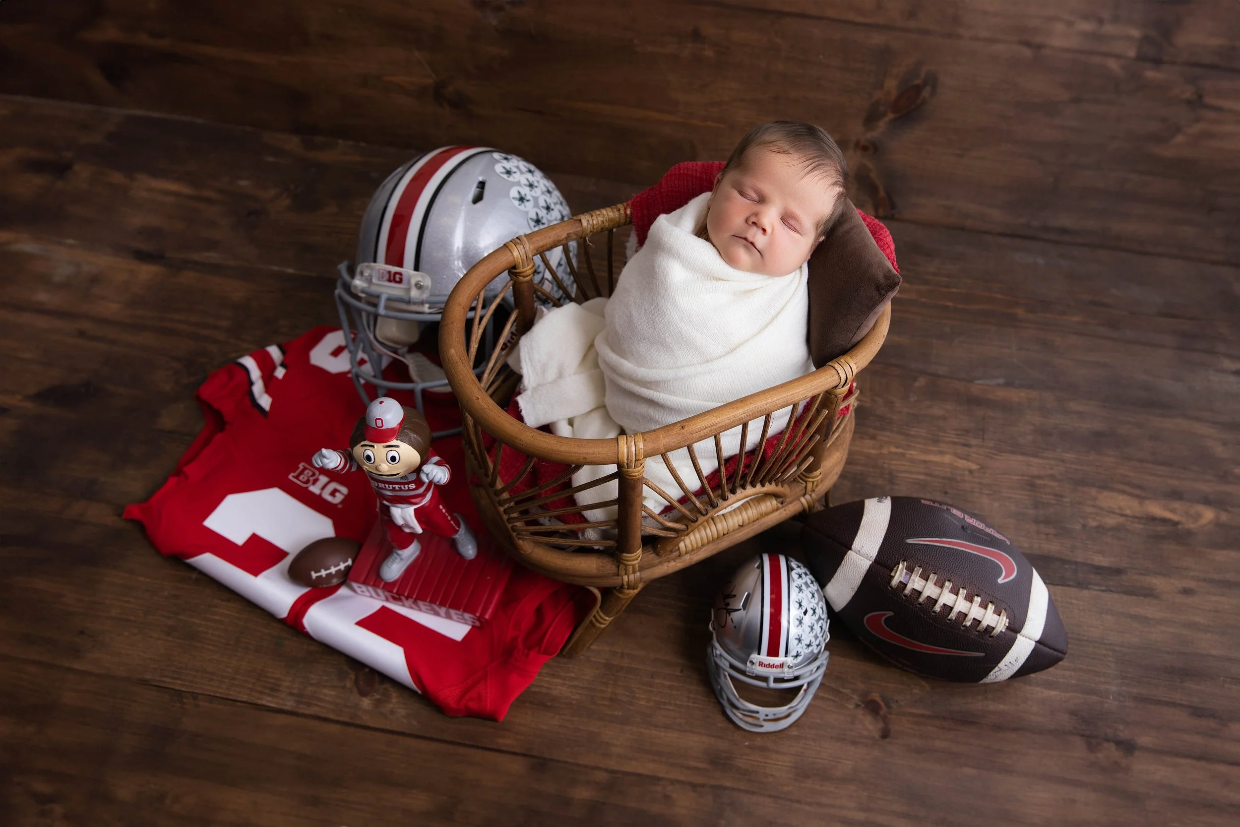 A sleeping baby wrapped in a white blanket inside a wicker basket surrounded by Ohio State University football memorabilia, including helmets, a jersey, a football, a toy figurine, and a small football on a wooden floor.