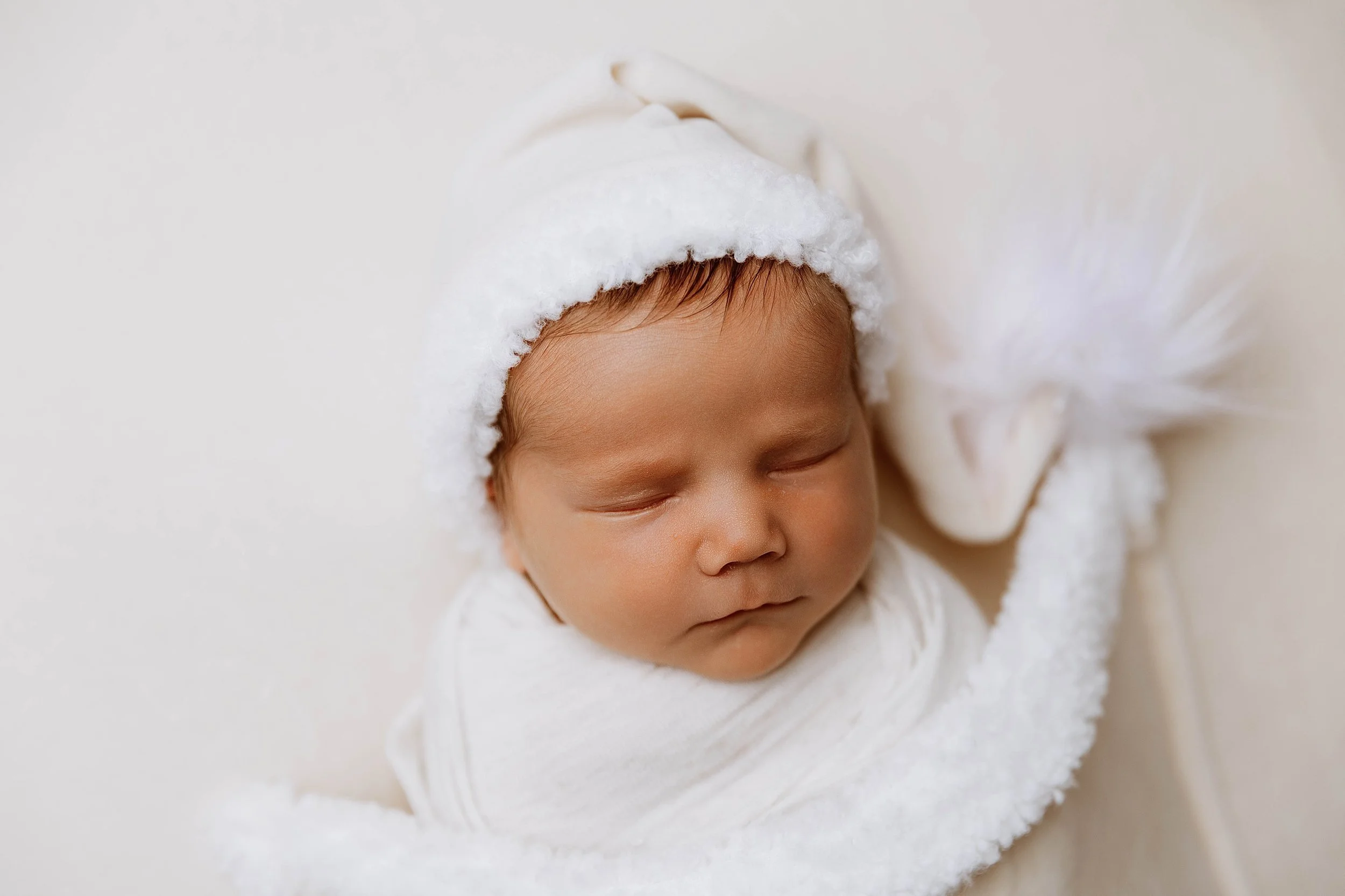 A sleeping newborn baby wearing a white Santa hat and wrapped in a white blanket with a fluffy white stuffed animal.