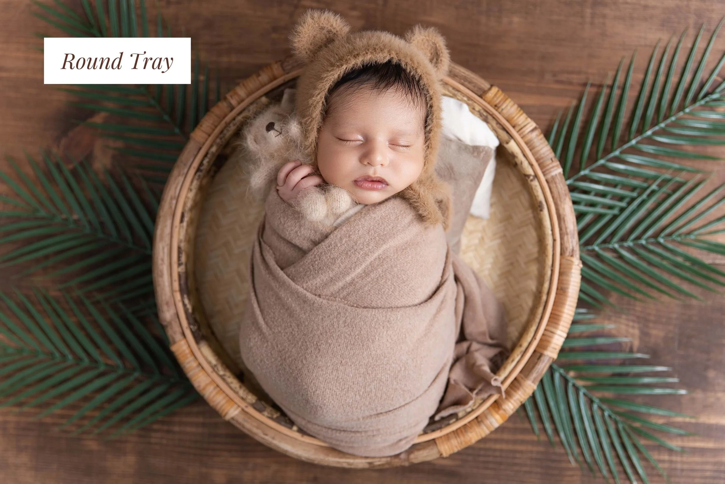 A sleeping baby wrapped in a beige blanket, wearing a bear hat, in a round wicker basket on a wooden surface with green palm leaves around it.