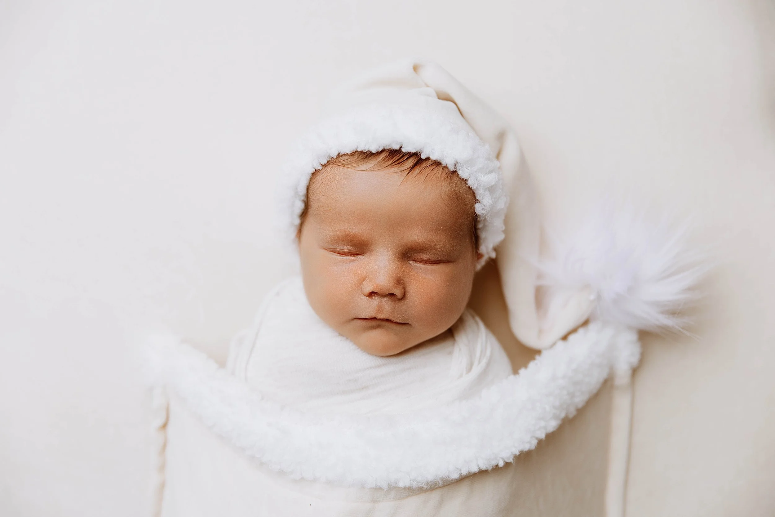 A sleeping baby wearing a white Santa hat and wrapped in a soft white blanket.