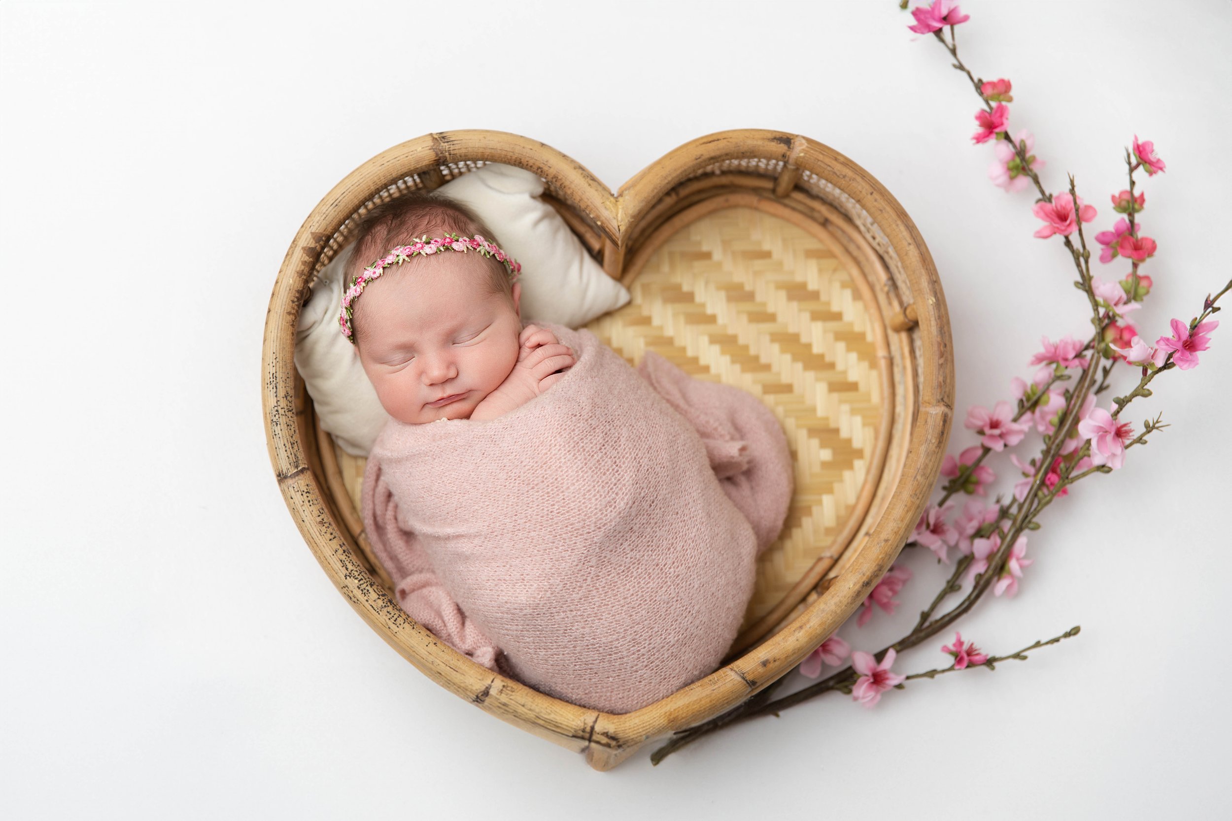 A sleeping newborn baby wrapped in a pink blanket, wearing a floral headband, inside a heart-shaped wicker basket, with pink cherry blossom branches nearby on a white background.