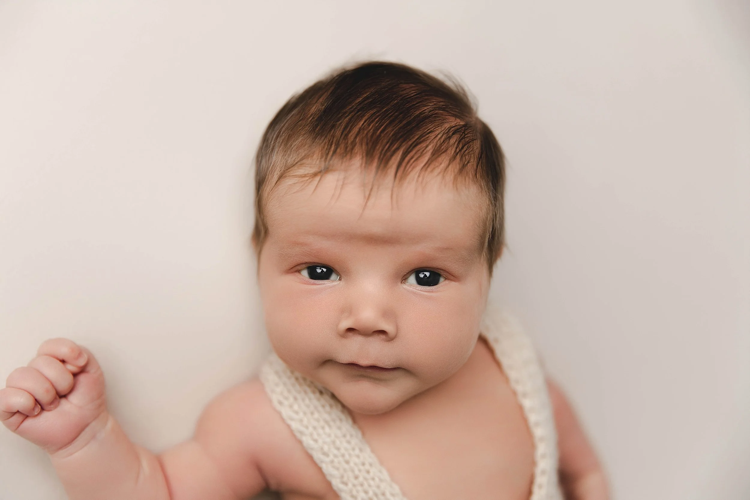 Close-up of a baby with dark hair, lying on a white surface, wearing a cream-colored knitted outfit, looking at the camera with a slight smile.
