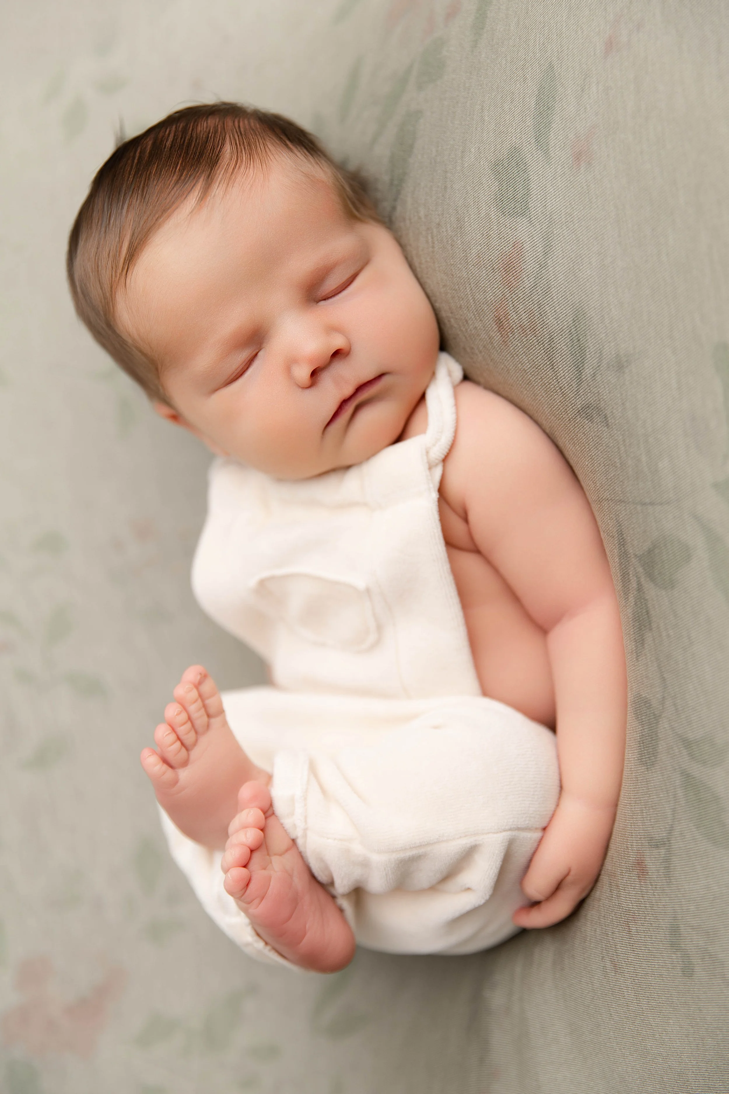 A sleeping baby with short brown hair, dressed in a white sleeveless outfit, lying on a soft surface with a floral pattern