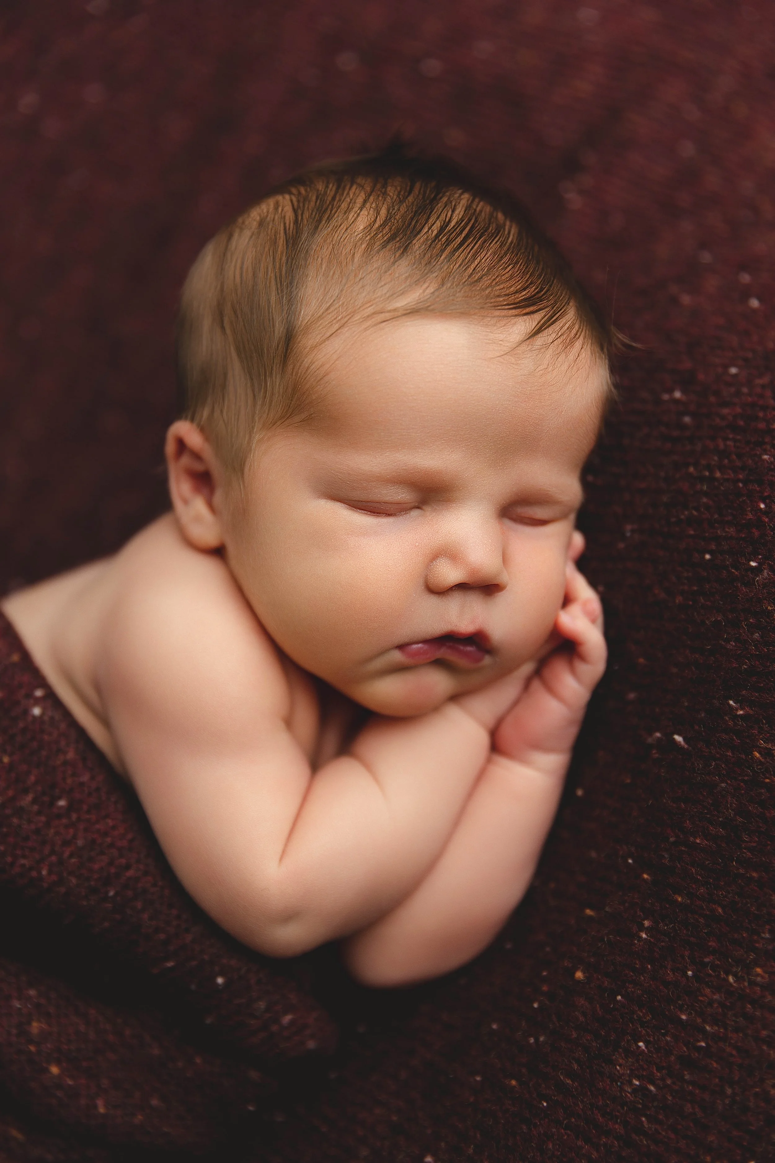 A sleeping newborn baby with closed eyes, resting on a soft, dark reddish-brown blanket, with tiny hands gently cupping the face.
