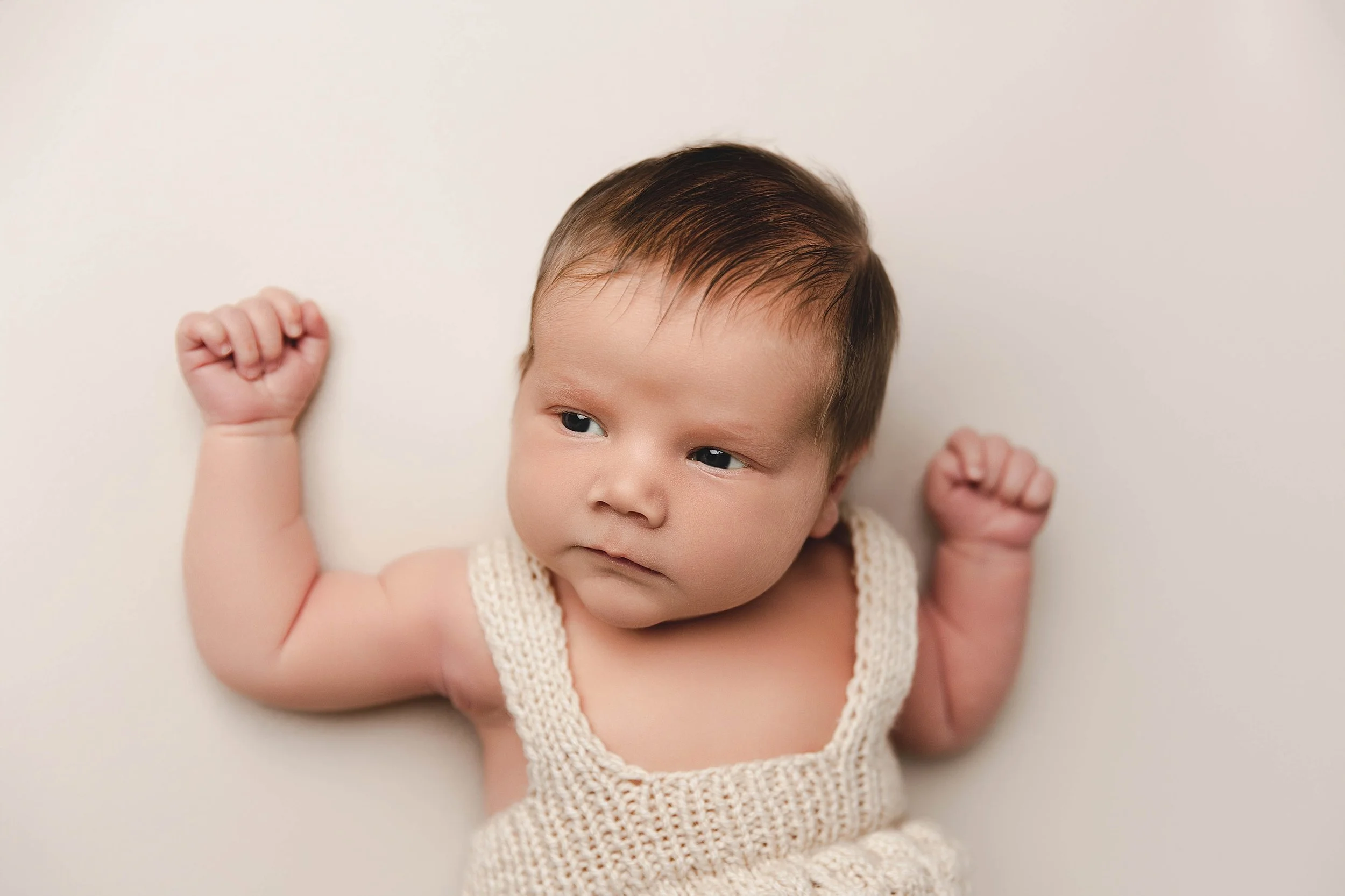 A baby lying on a white surface, wearing a knitted white outfit, with arms raised and a thoughtful expression.