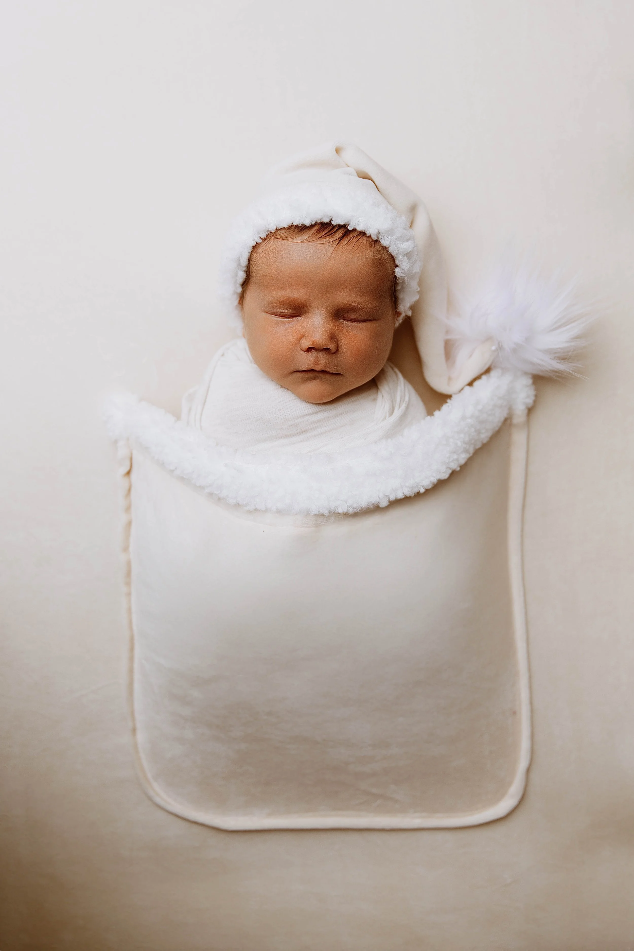 A sleeping baby wearing a white hat with a fluffy pom-pom and a white blanket, lying on a soft cream-colored surface.