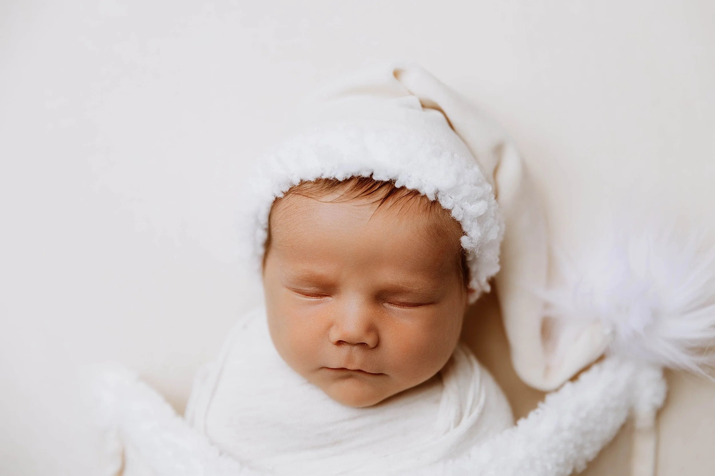 Close-up of a sleeping baby wearing a white hat with a fluffy brim, wrapped in a white blanket, lying on a soft white surface.