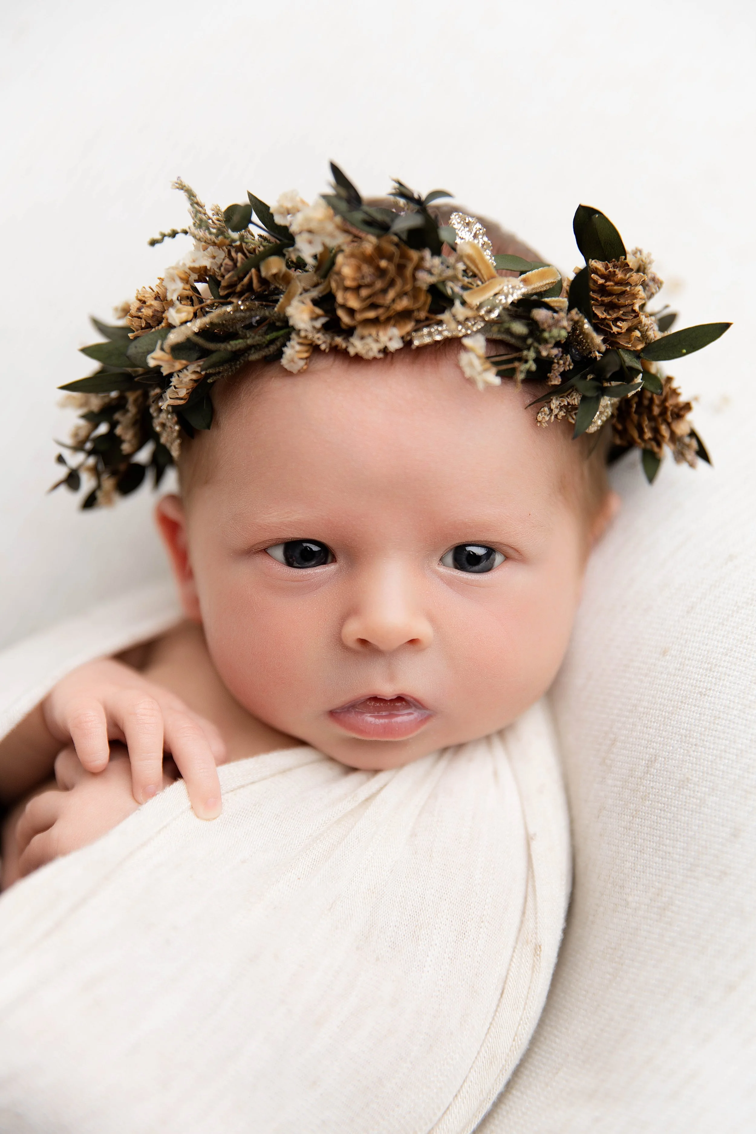 Close-up of a baby with blue eyes, wearing a cream outfit and a floral crown made of pinecones and leaves, lying on a white surface.
