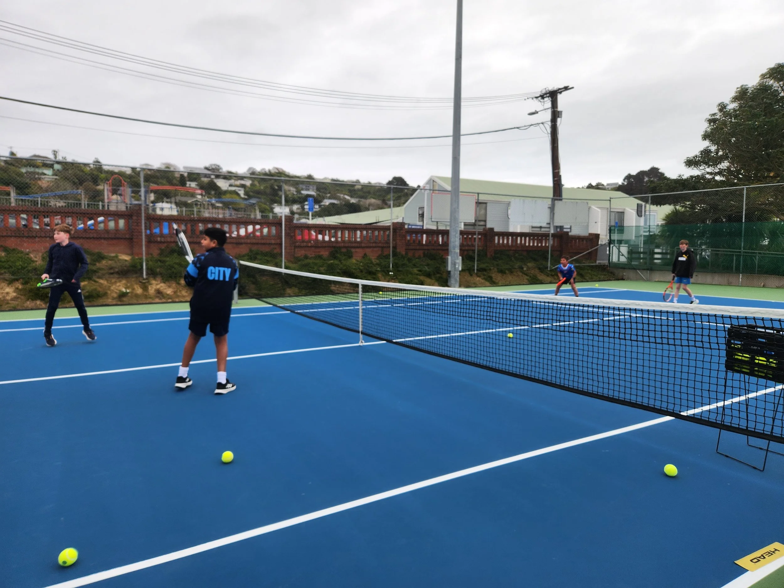 Four kids playing tennis on an outdoor blue tennis court with a black net, tennis balls, and a basket of tennis balls on the right side. The background shows trees, a fence, and some buildings under cloudy skies.