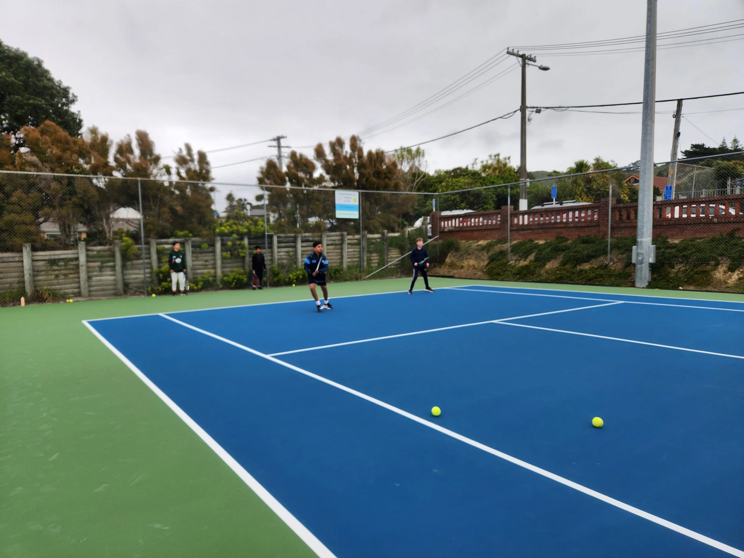 People playing tennis on an outdoor court on a cloudy day, with several tennis balls on the ground, a tall chain-link fence, and trees in the background.
