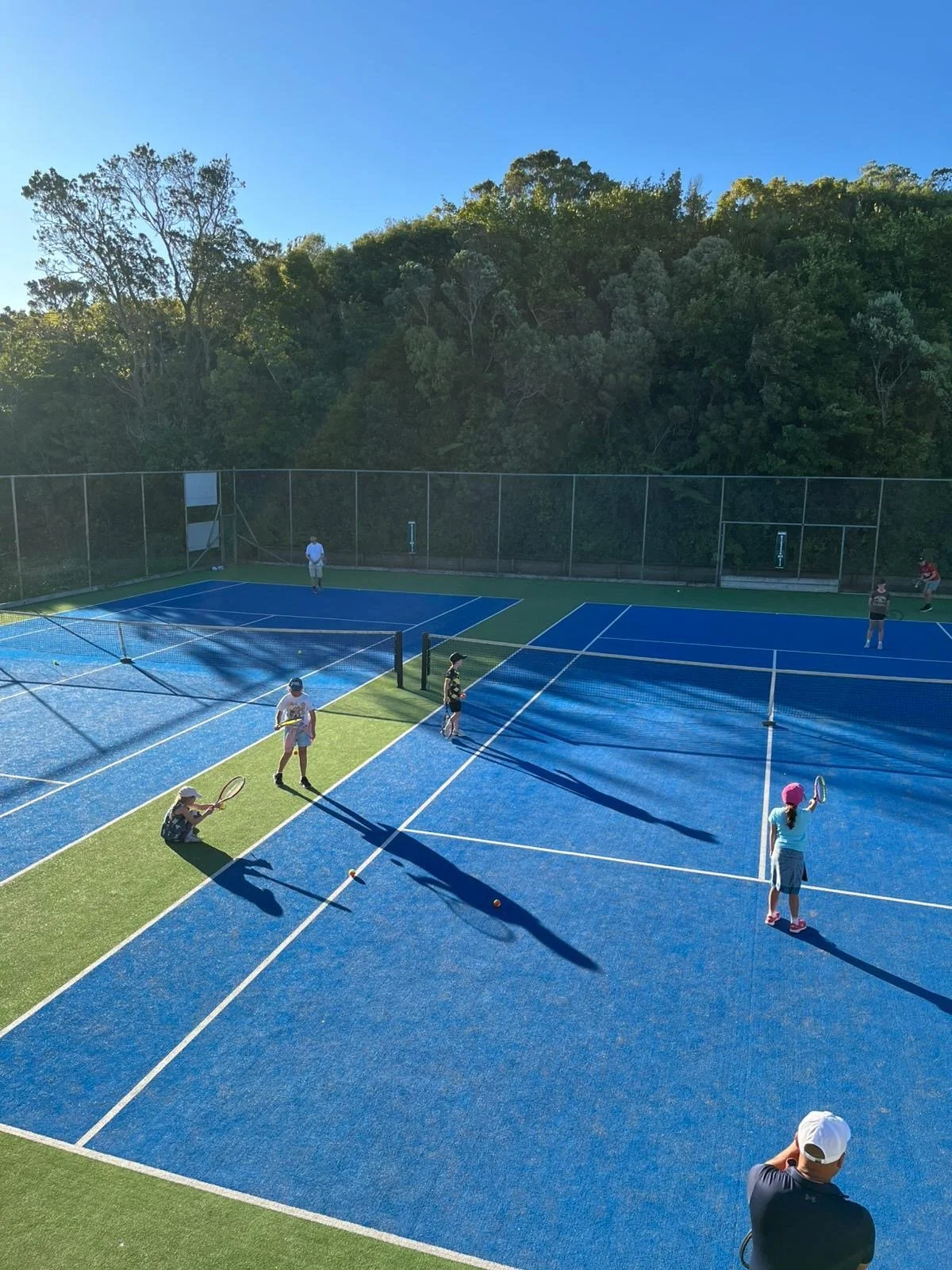 Children playing pick-up tennis on bright blue courts with a parent watching and lush green trees in the background on a sunny day.