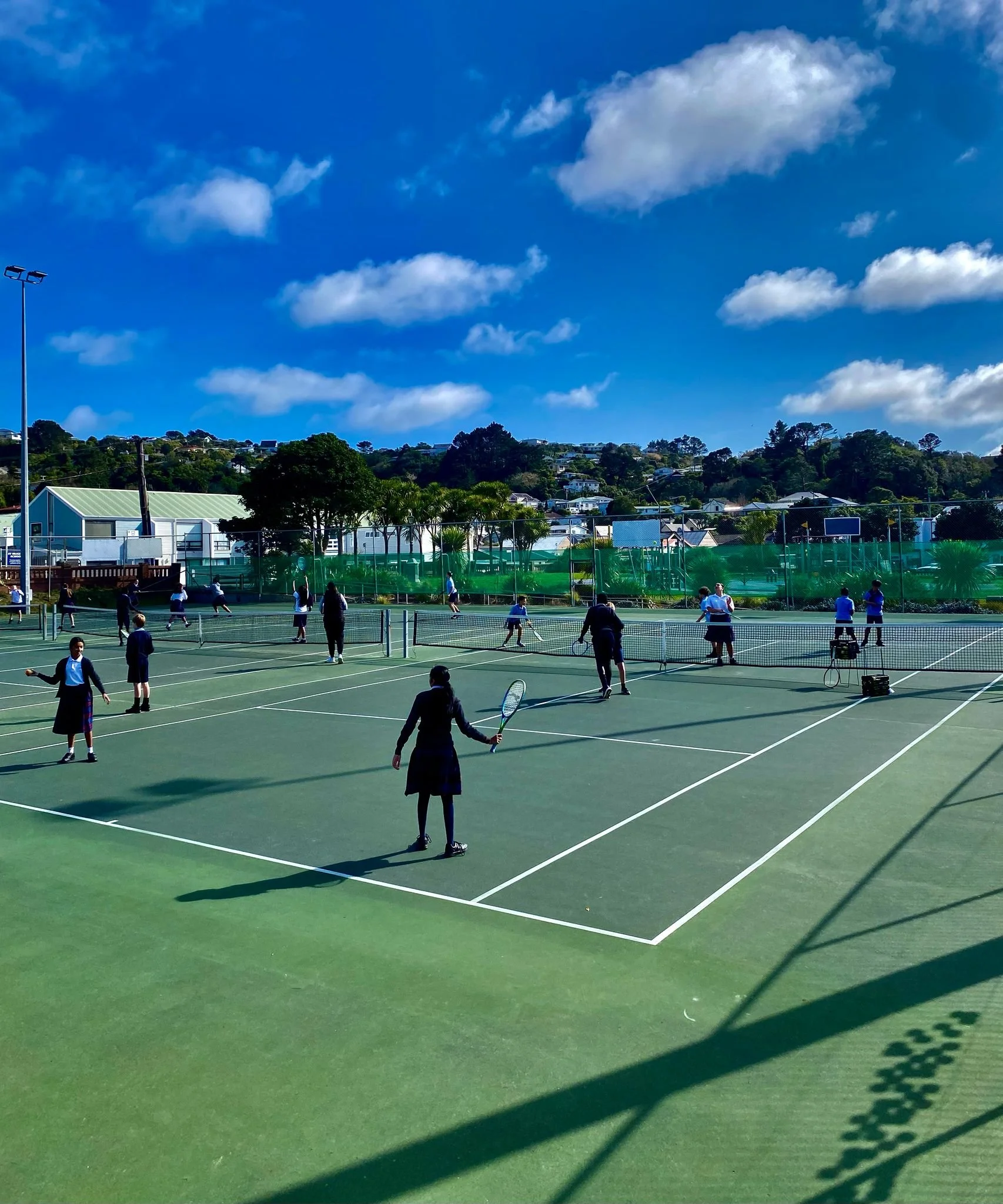 Multiple people, including children and adults, playing tennis on a sunny outdoor tennis court with a green surface and a backdrop of trees and buildings under a blue sky with fluffy clouds.