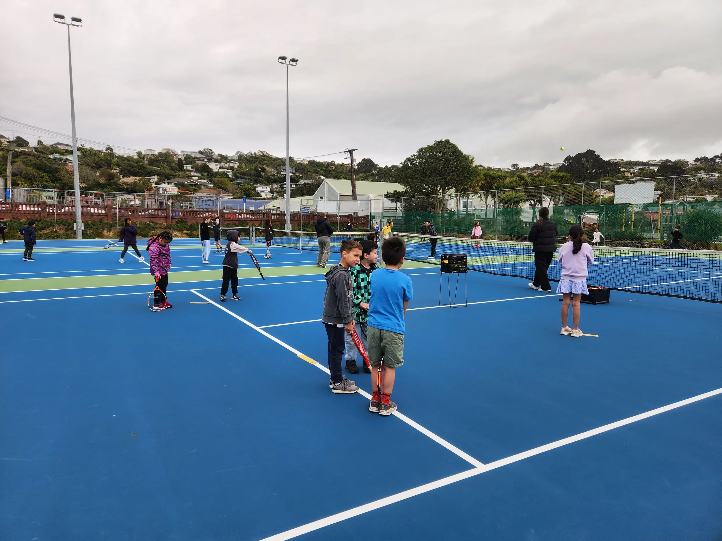 Children and adults playing and practicing tennis on blue and green outdoor tennis courts with net and equipment, set against a backdrop of houses, trees, and cloudy sky.