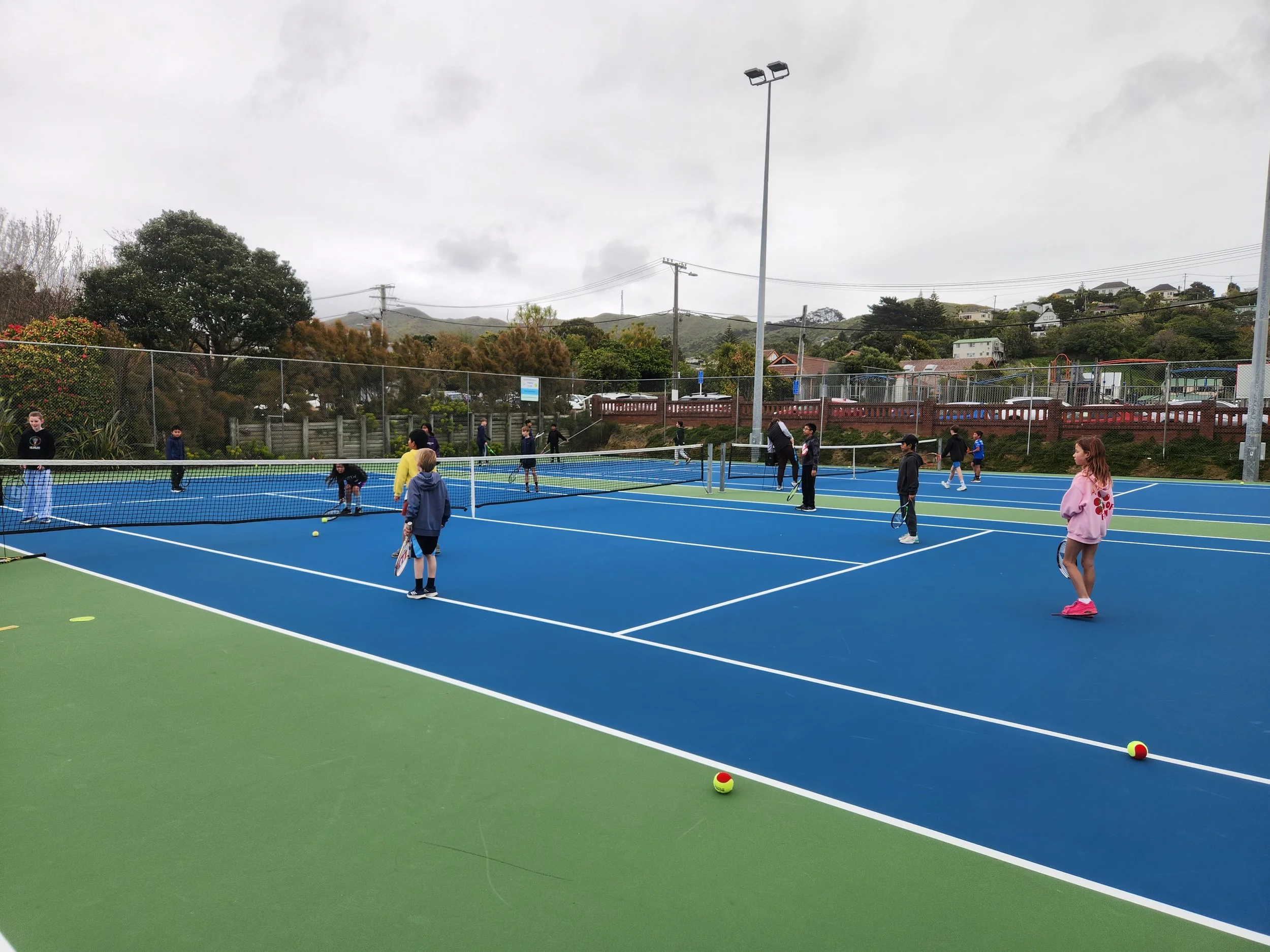Children and adults playing tennis on blue and green outdoor courts, with hills and houses in the background under cloudy skies.