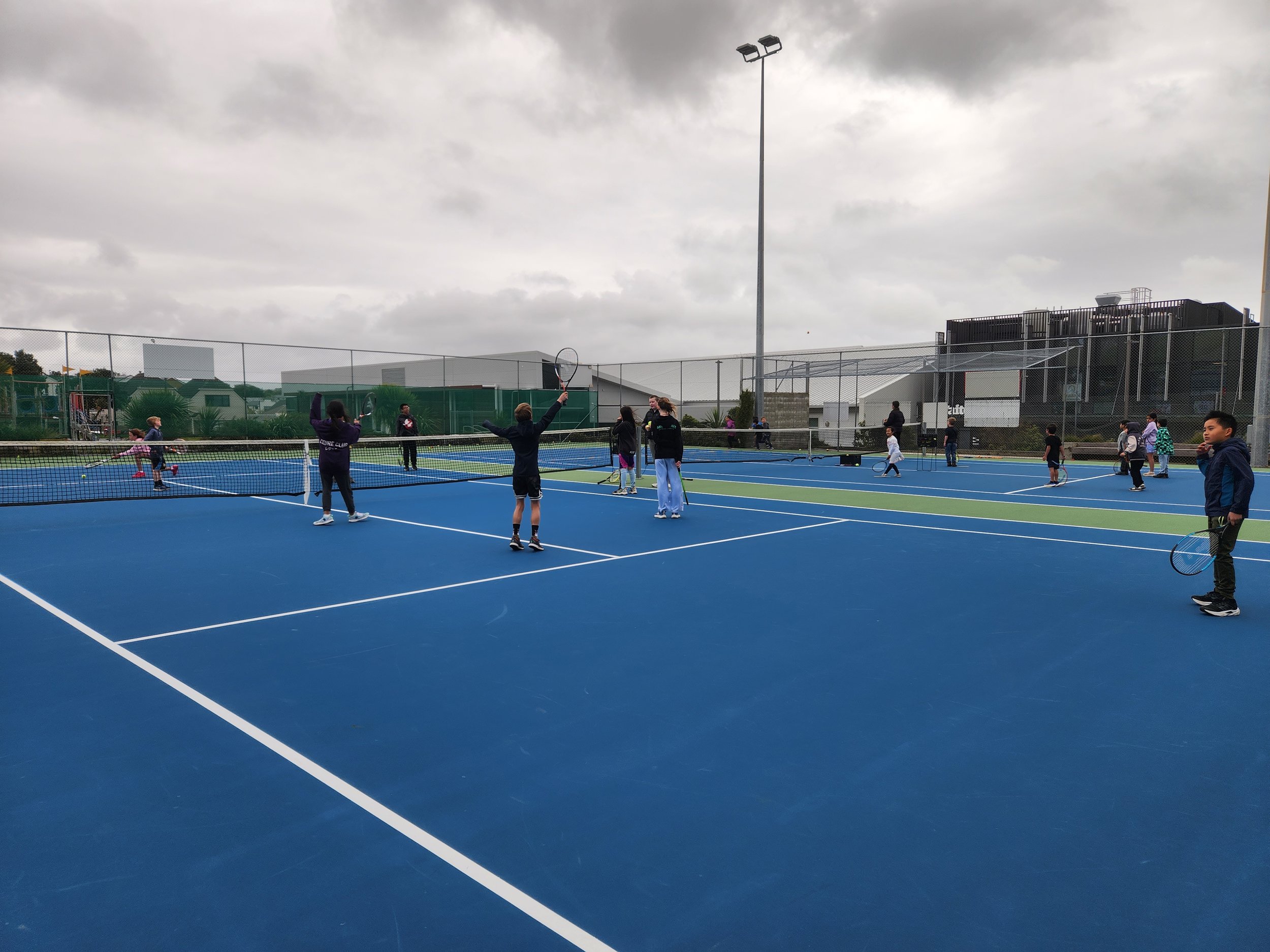 People playing tennis on an outdoor court with blue surface, overcast sky, and fenced surroundings.