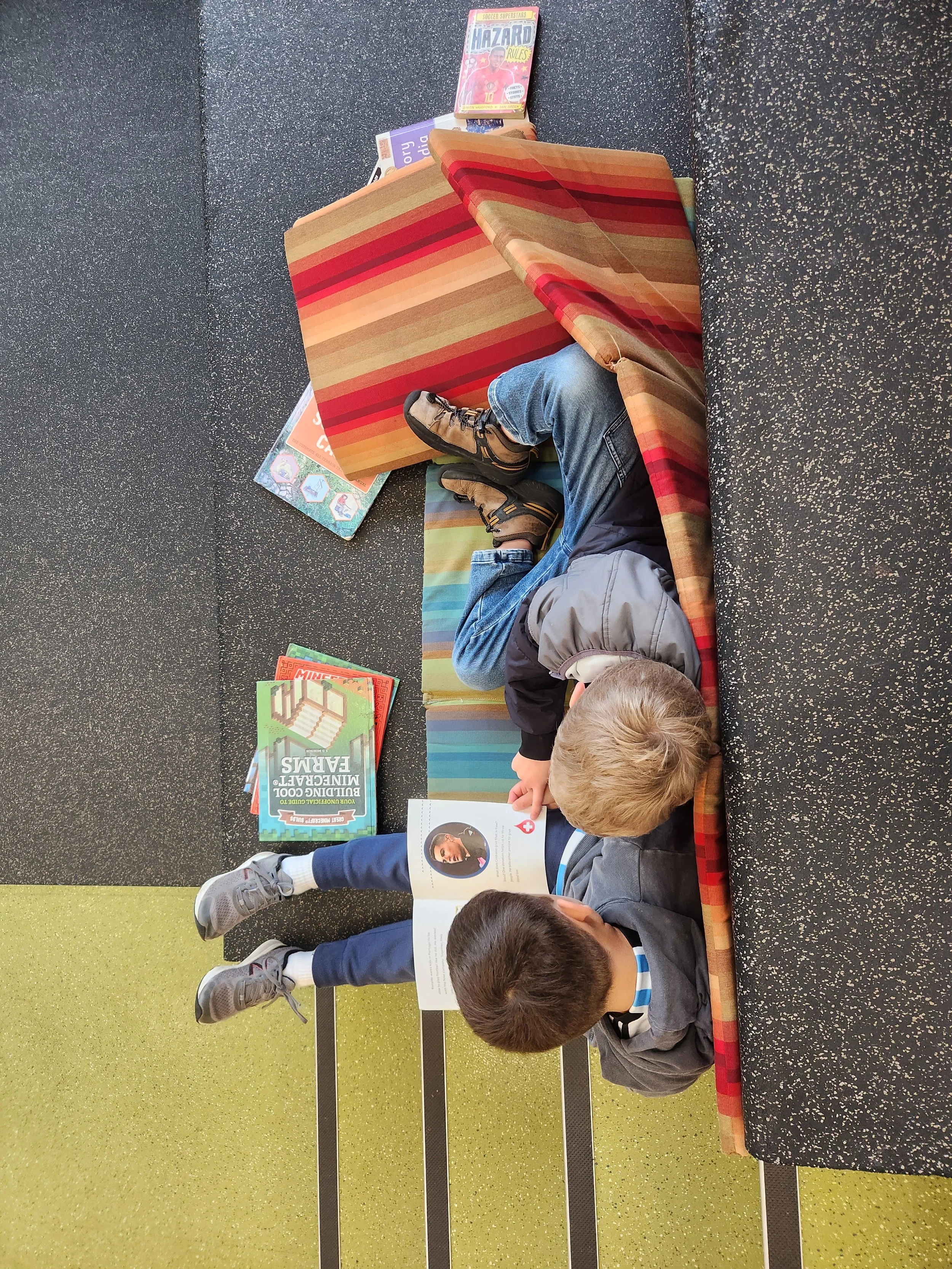 Two young boys reading books while lying on a floor with a black and green speckled surface. There are books on the floor and a striped, multicolored couch or bench behind them.
