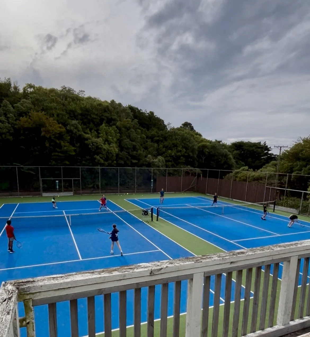 People playing tennis on outdoor blue tennis courts, surrounded by green trees and a cloudy sky.
