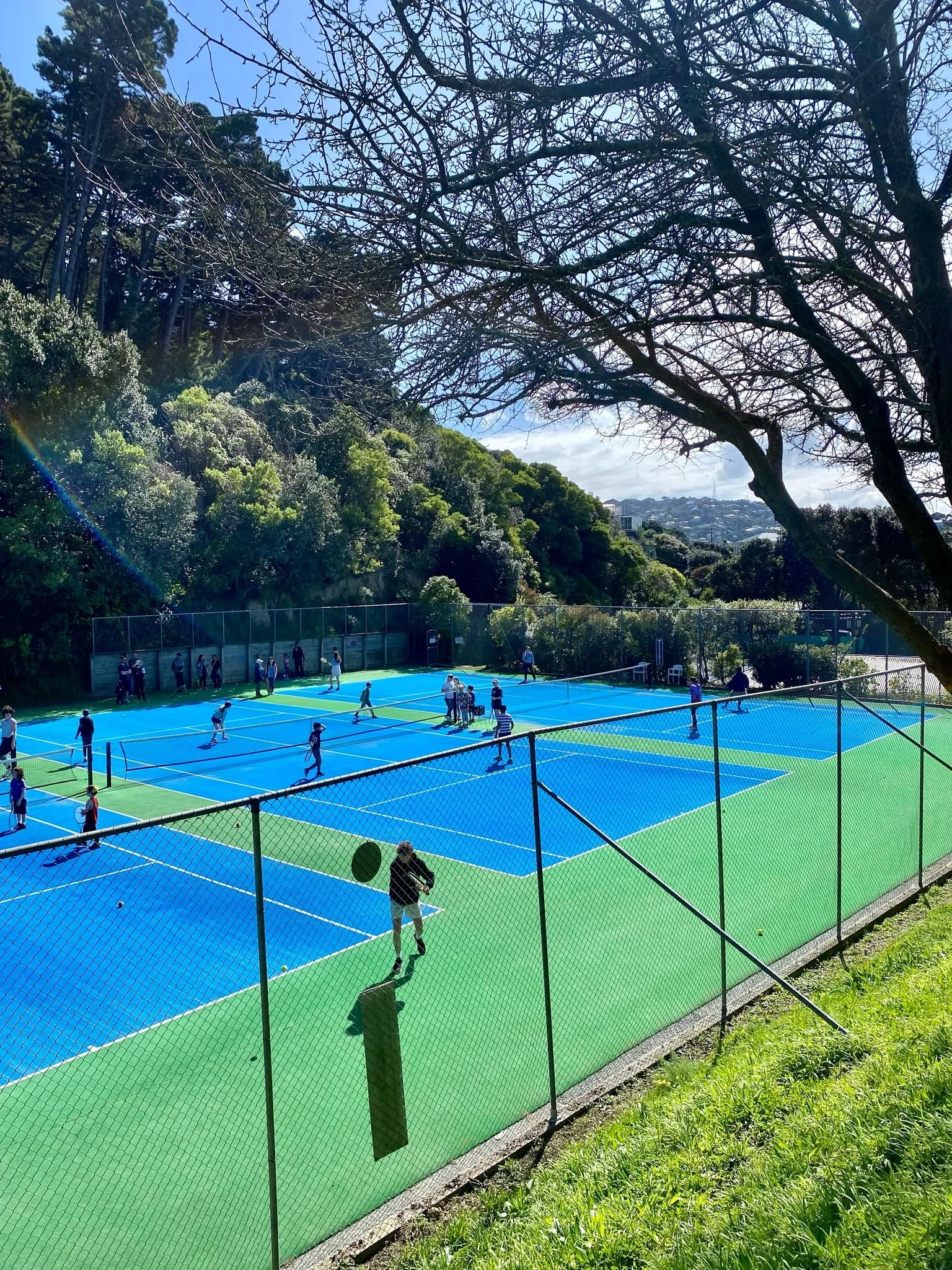 Tennis courts with people playing and practicing tennis under a sunny sky, surrounded by trees and a hillside.