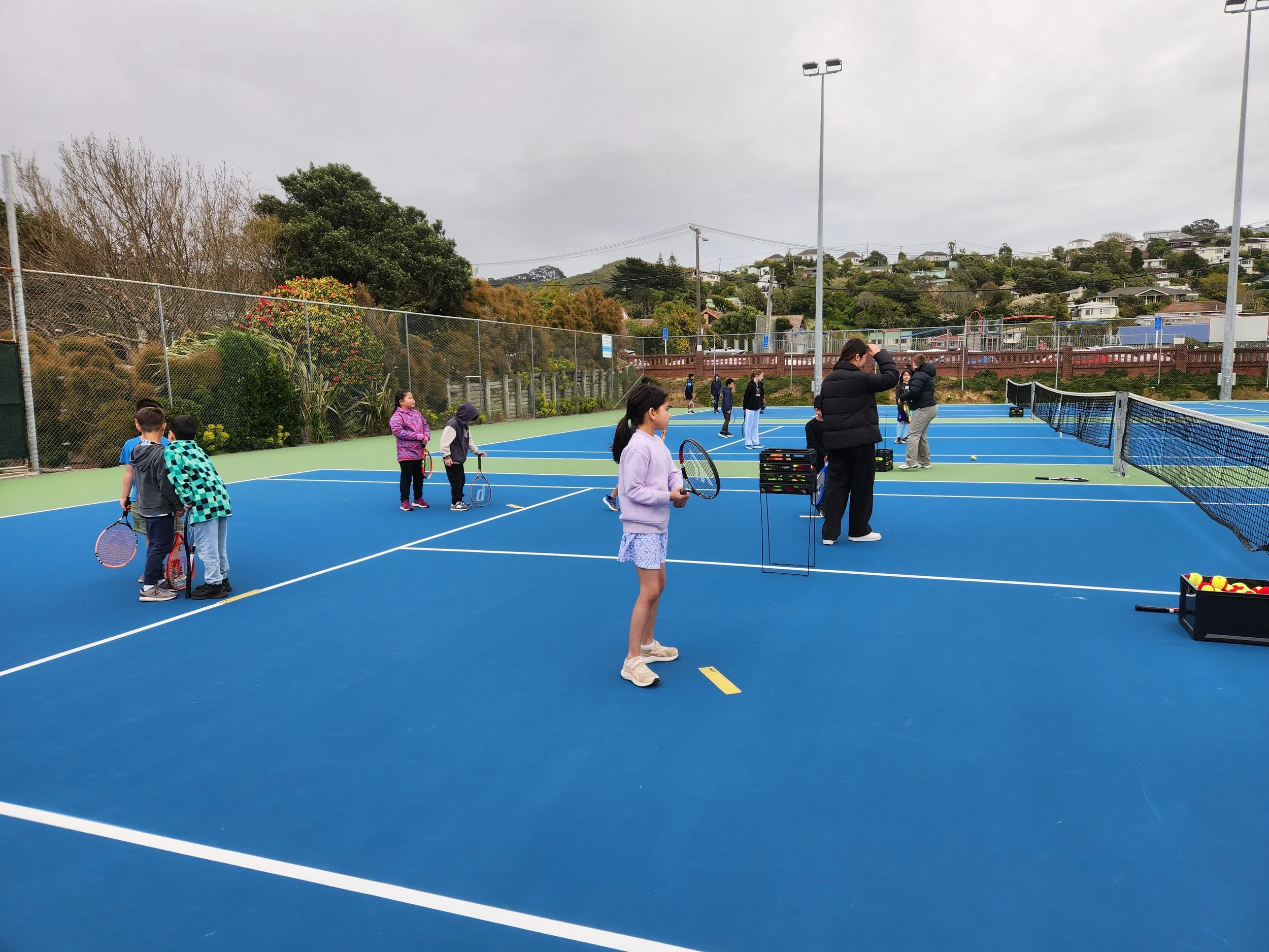 Children playing tennis on an outdoor court with coaches and adults, surrounded by trees and hillside houses under a cloudy sky.