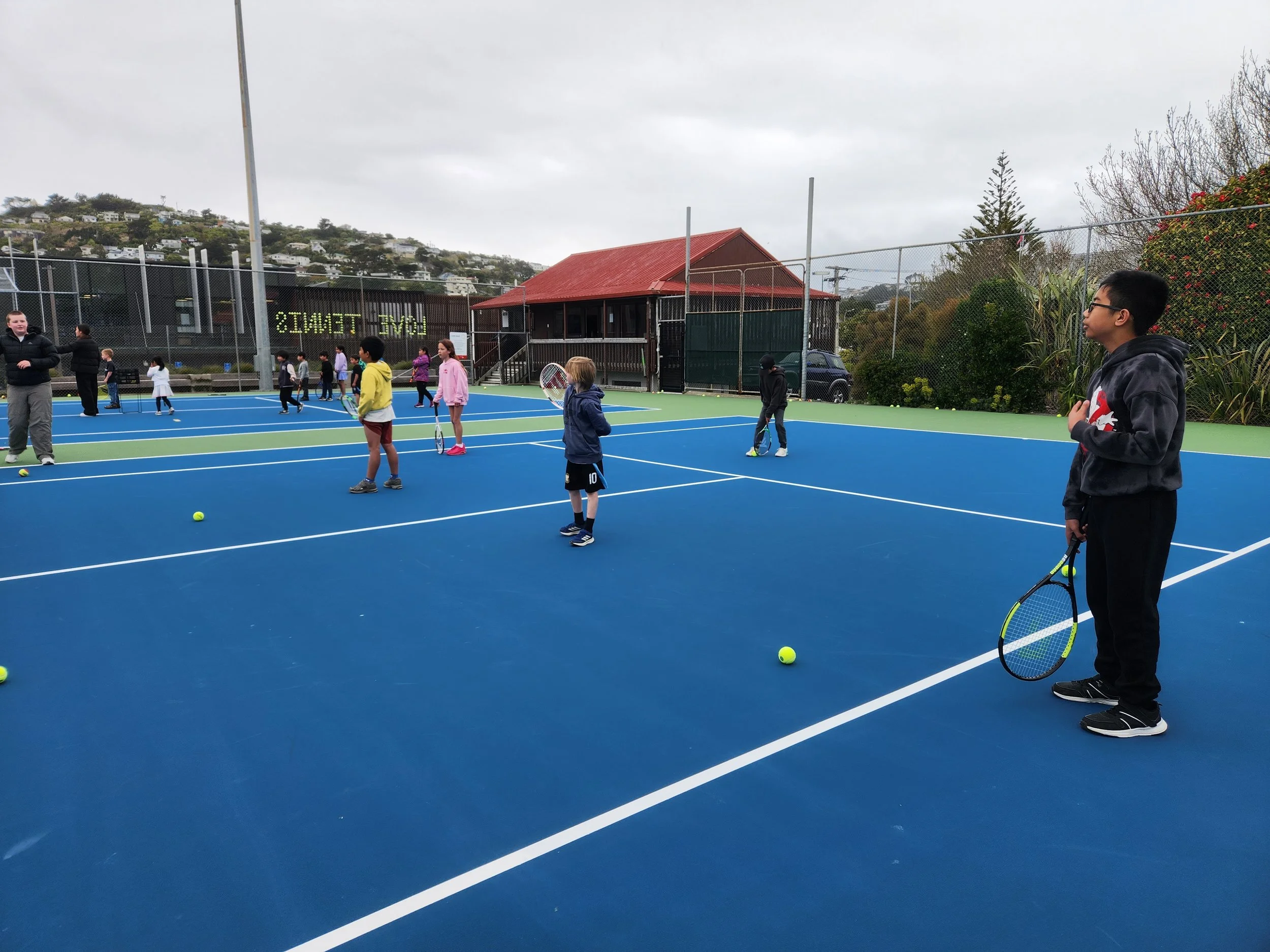 Children practicing tennis on a blue tennis court with an instructor, holding tennis rackets and yellow tennis balls, under an overcast sky.