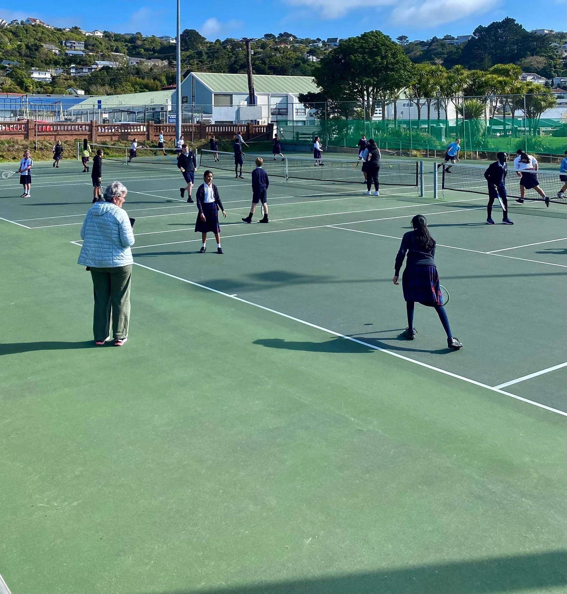 School students playing tennis on an outdoor court on a sunny day, with a teacher or coach overseeing the activity.