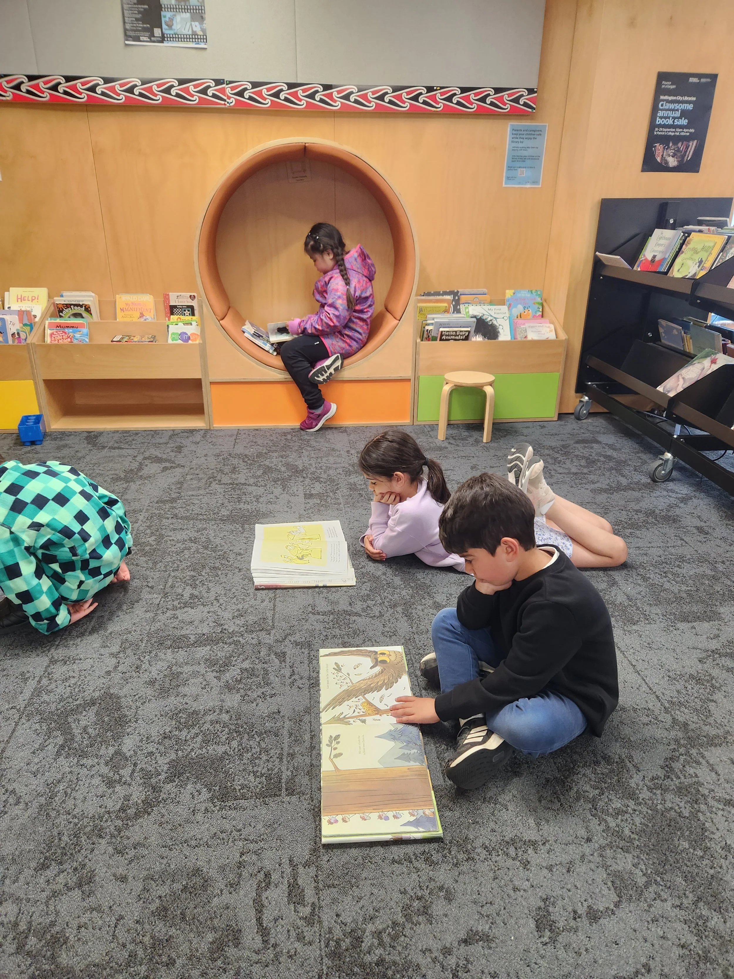 Four children reading books on the carpeted floor of a library. One girl seated in a circular nook reading a book, three kids lying or kneeling nearby with books open in front of them.