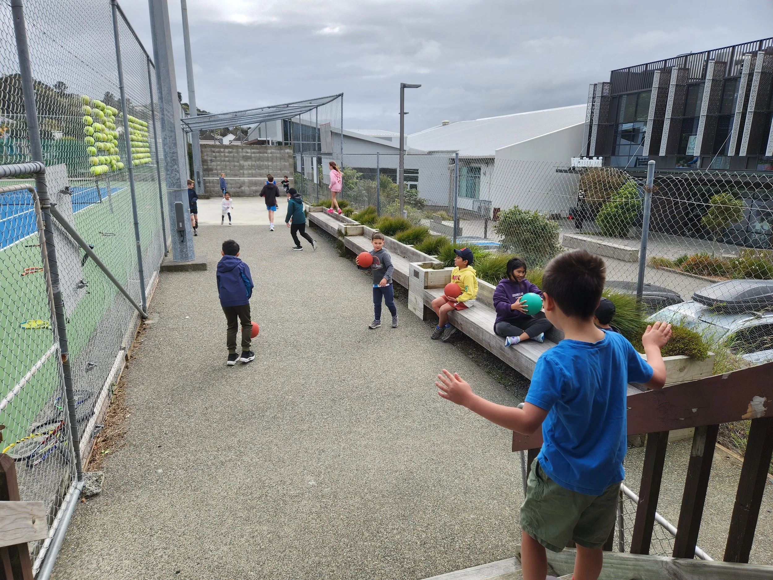 Children playing with balls in a school yard enclosed by a chain-link fence, with some sitting on a wooden bench and others running around.